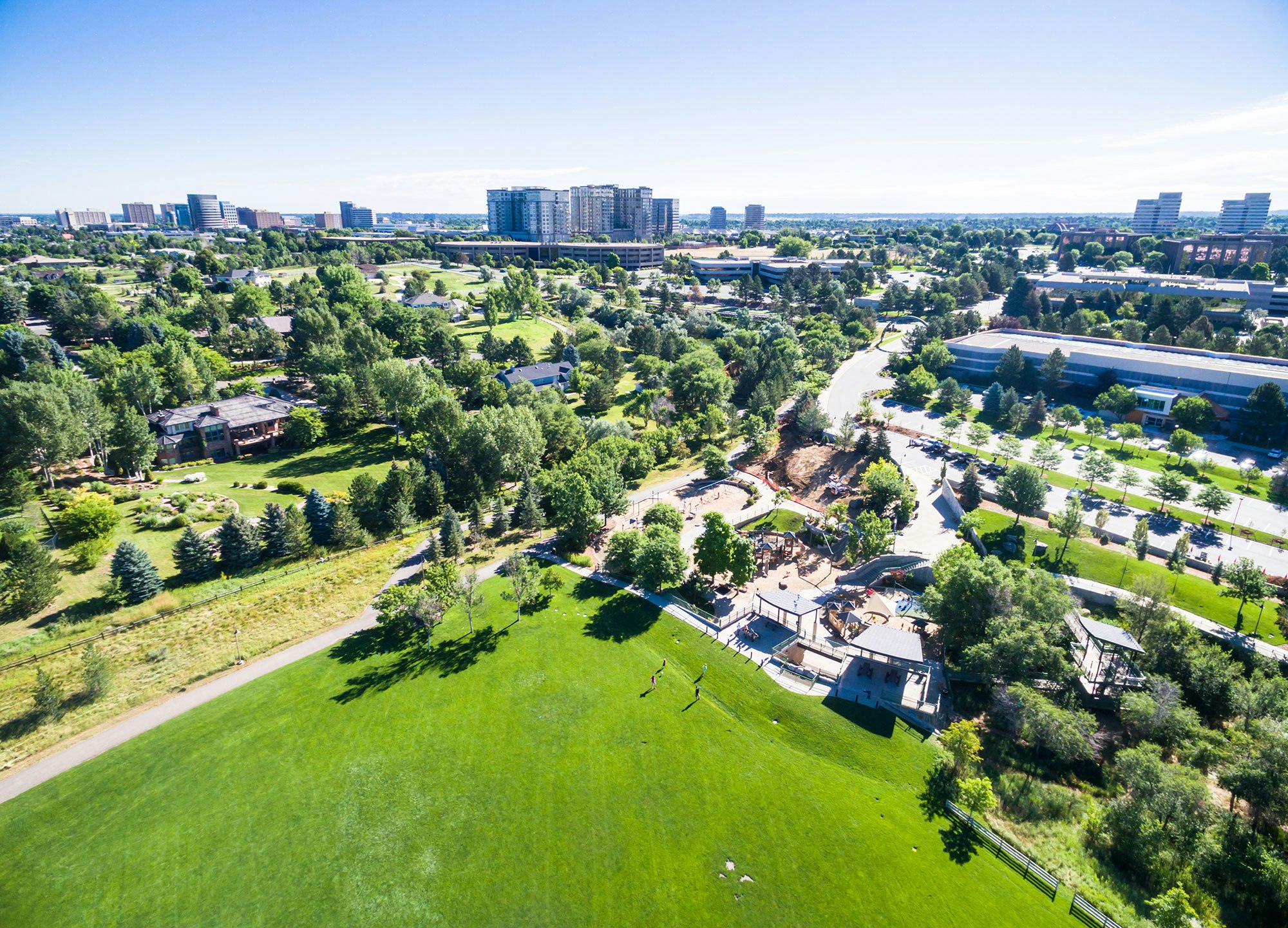 Aerial view of a lush green park with walking paths, playground and nearby residential area set against an urban skyline.