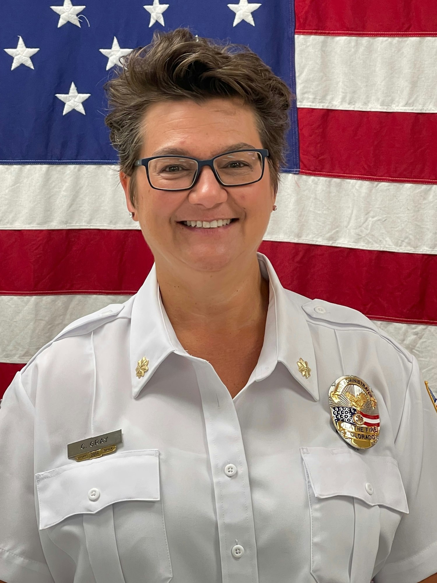 Christine Gray in a white uniform with a badge, in front of an American flag.