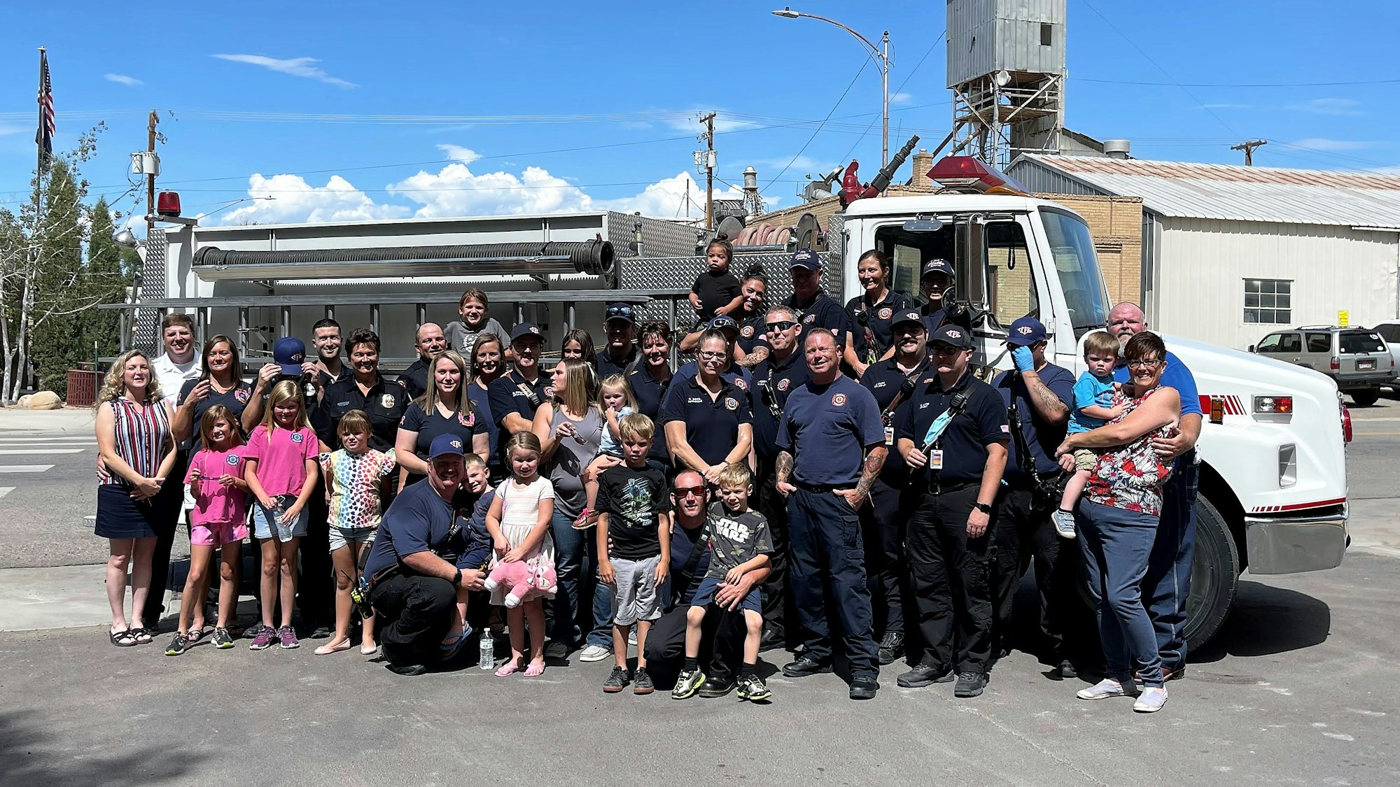 A large group of firefighters and families poses together in front of a fire truck on a sunny day.