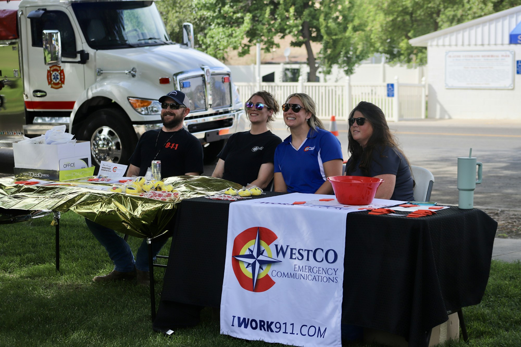 Four people behind a table with a "Westco Emergency Communications" banner, promotional materials, and a fire truck in the background.