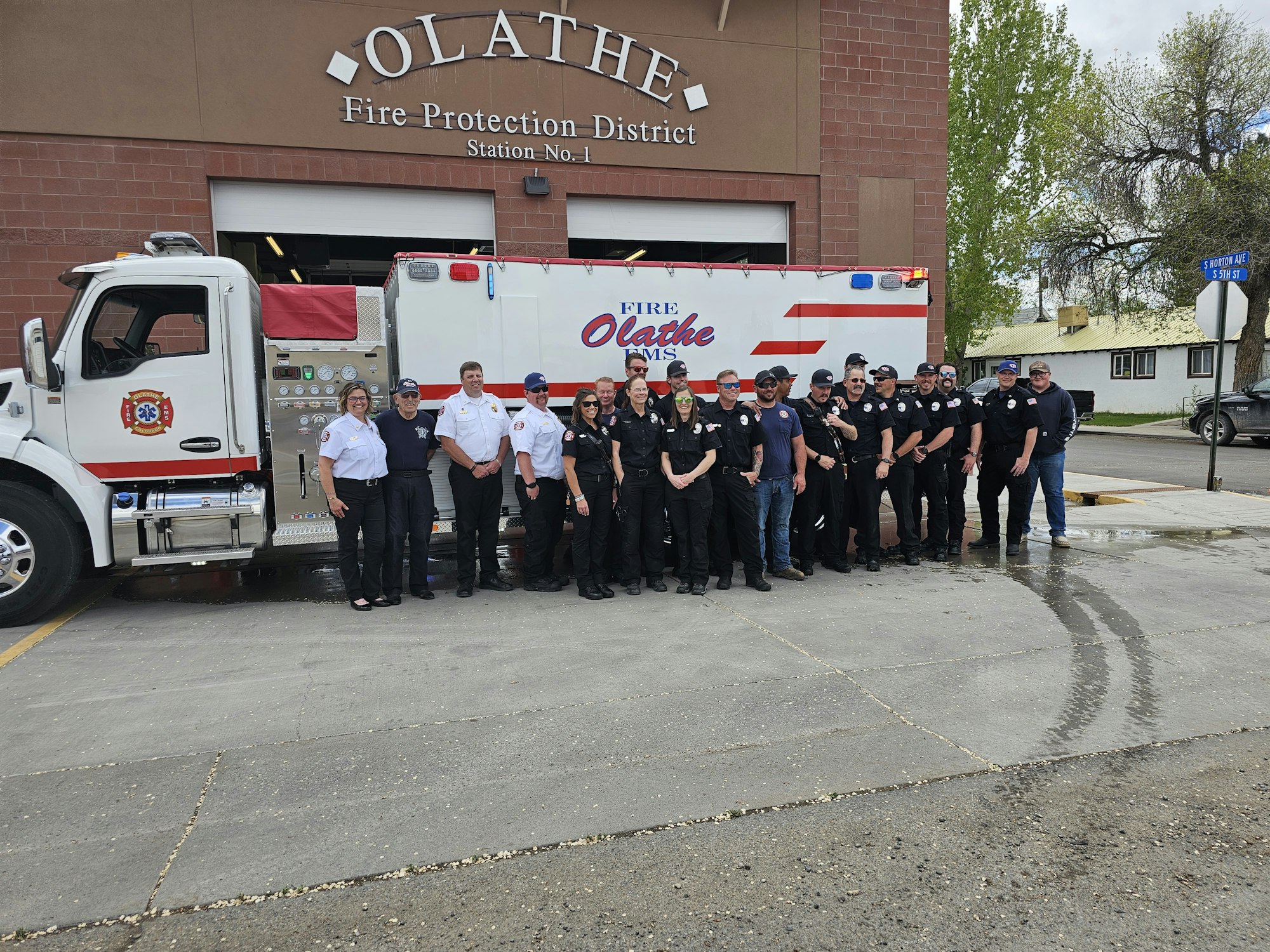 A group of firefighters and EMS personnel standing in front of a fire truck by their station.