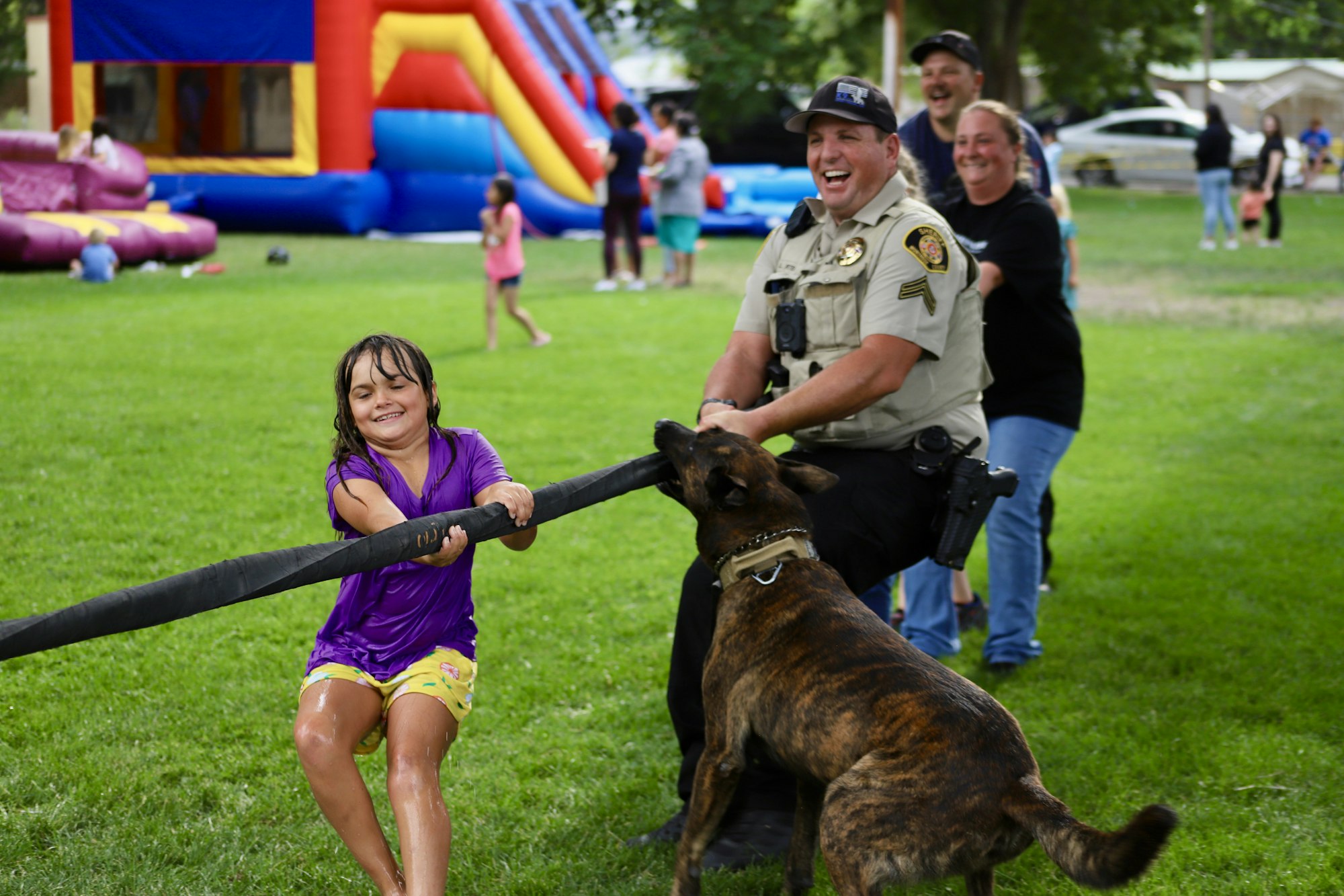 A girl plays tug-of-war with a police officer and his dog at a community event, with onlookers smiling in the background.
