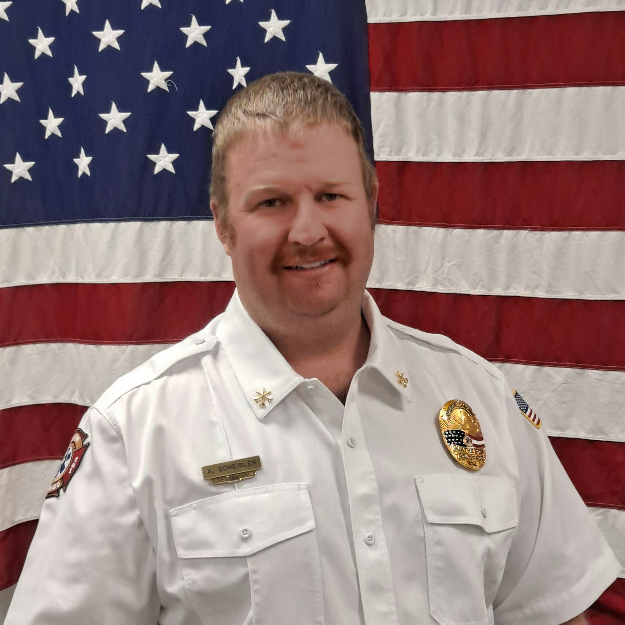 Person in a uniform with badges, tilted sideways, standing before an American flag.