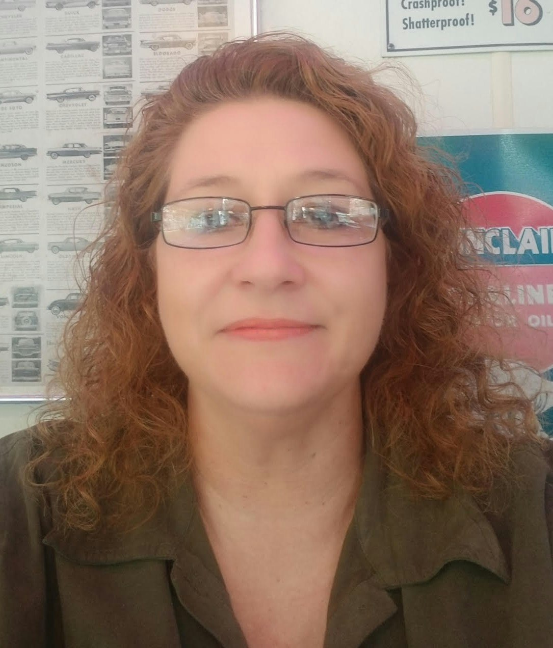 A woman with curly hair and glasses smiles for a selfie in front of a vintage car advertisement background.