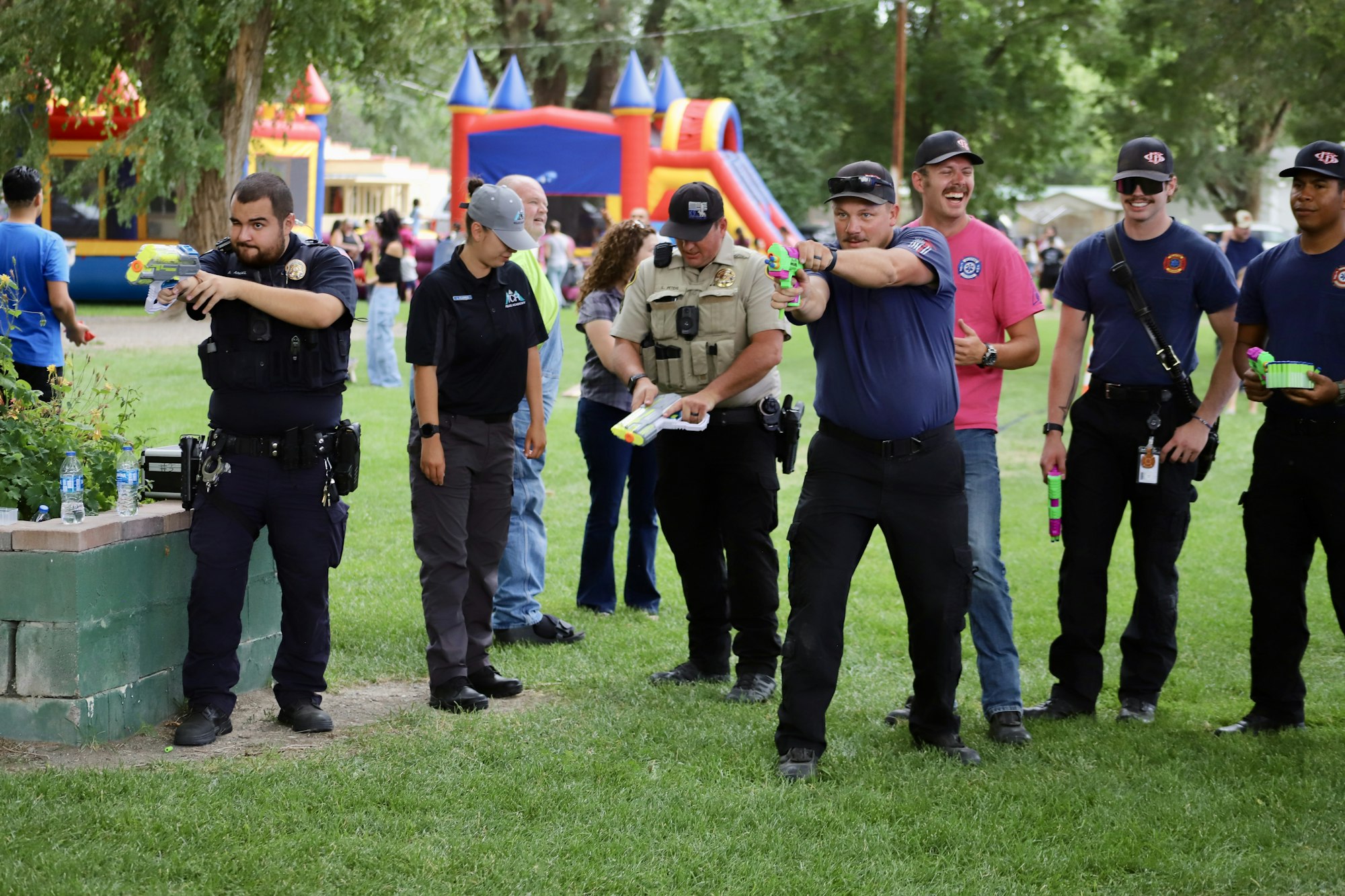 Police and firefighters with toy water guns, having fun, with inflatable bounce houses in the background.