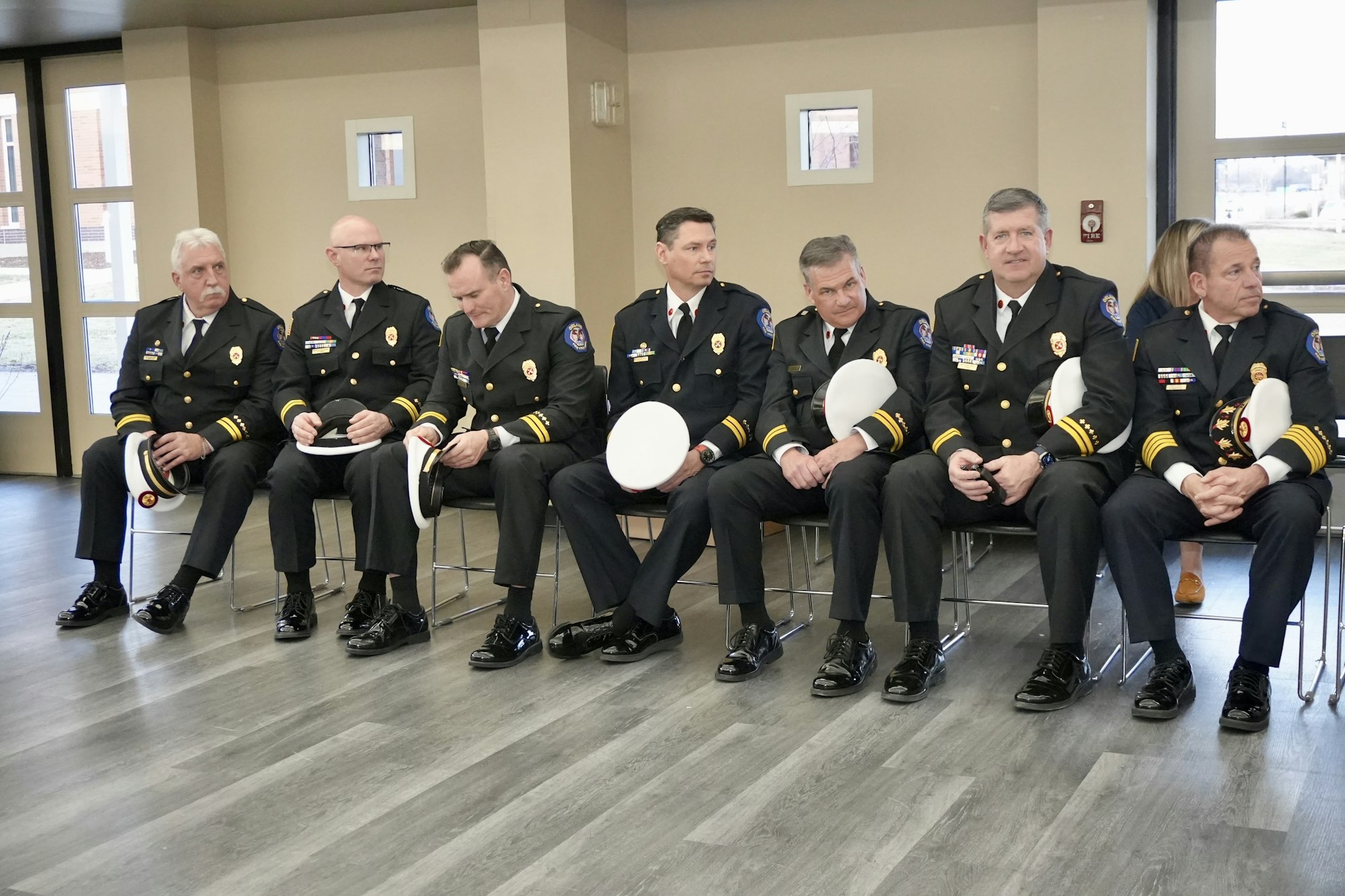 Seven uniformed officials sitting on chairs indoors, holding white hats.