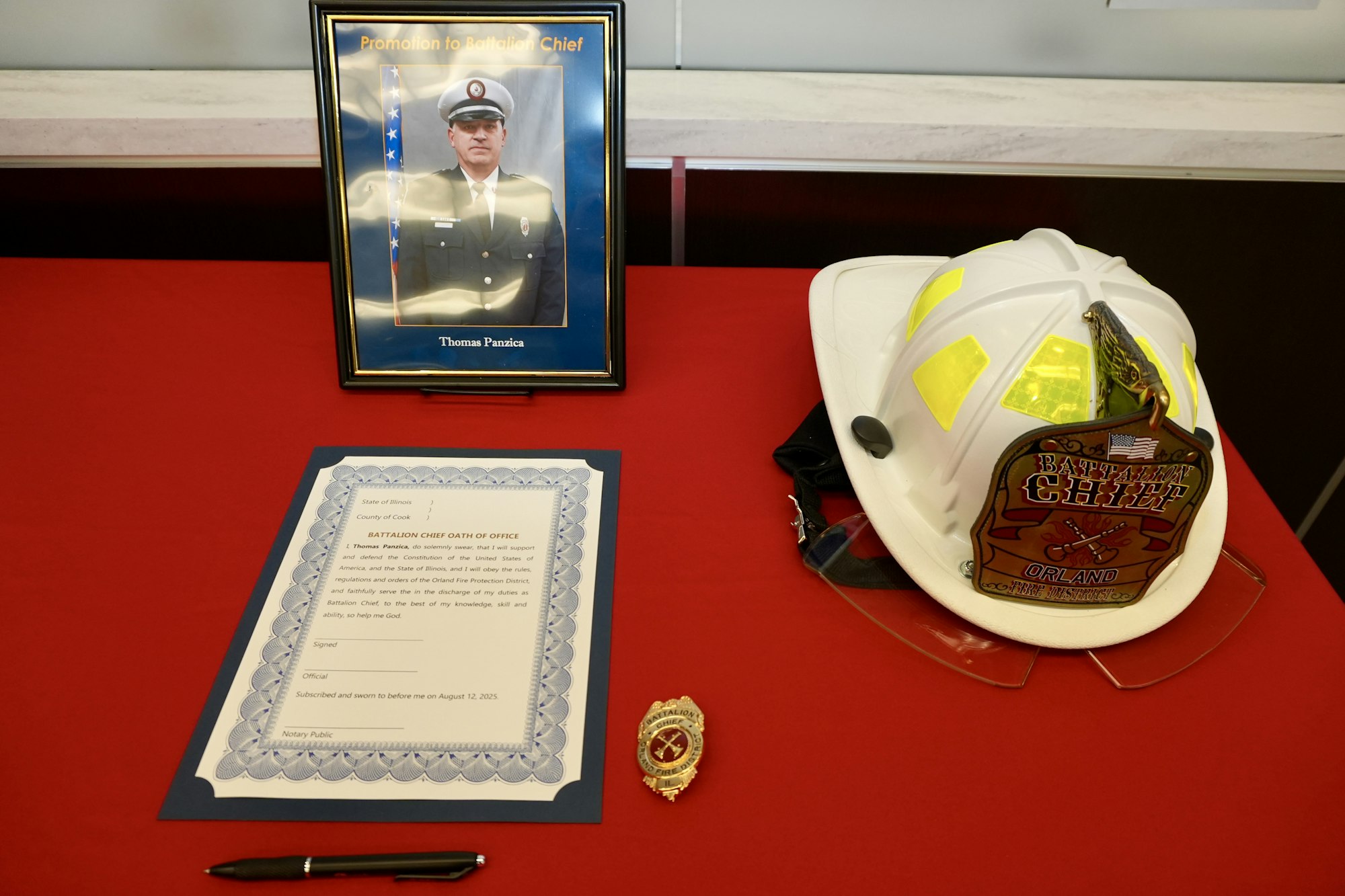 A framed photo, firefighter helmet, badge, and an oath document on a red tablecloth.