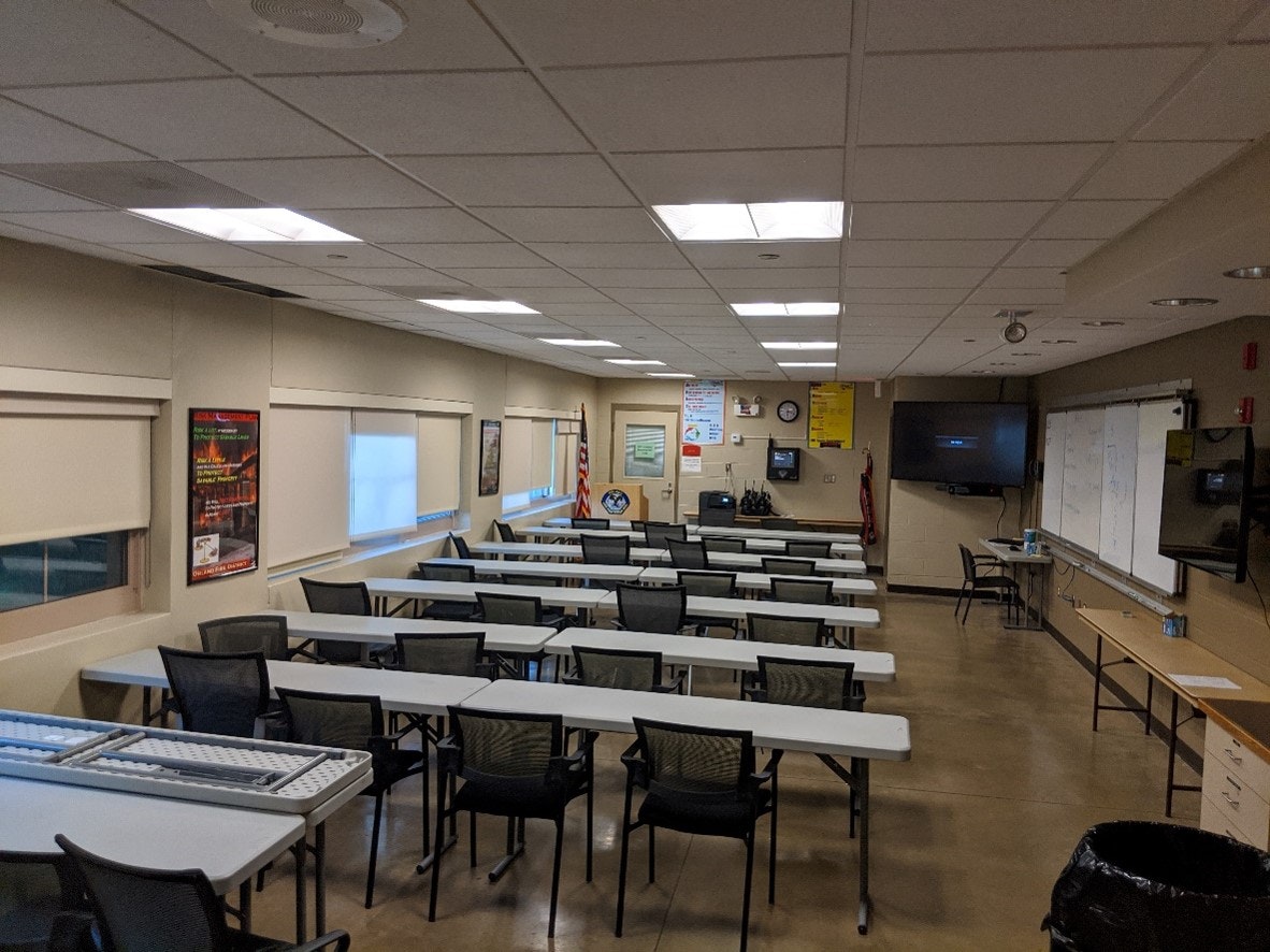 Empty classroom with rows of tables and chairs, whiteboards, projection screen, TV, and US flag.
