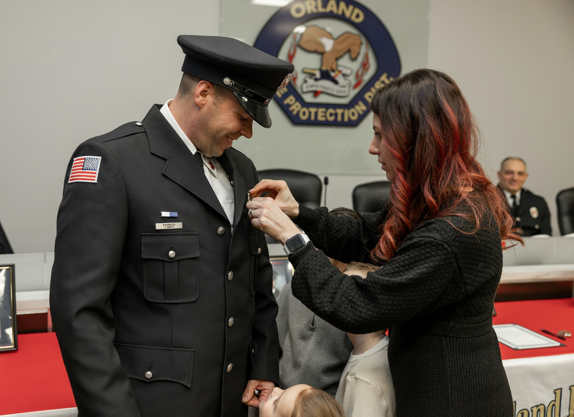 A woman pins a badge on a smiling man in uniform, while children watch, in a ceremony with an official backdrop.