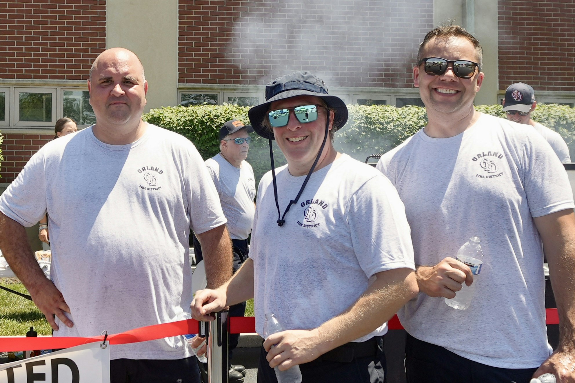 Three people smiling in matching shirts, holding water bottles, with others in the background at an outdoor event.