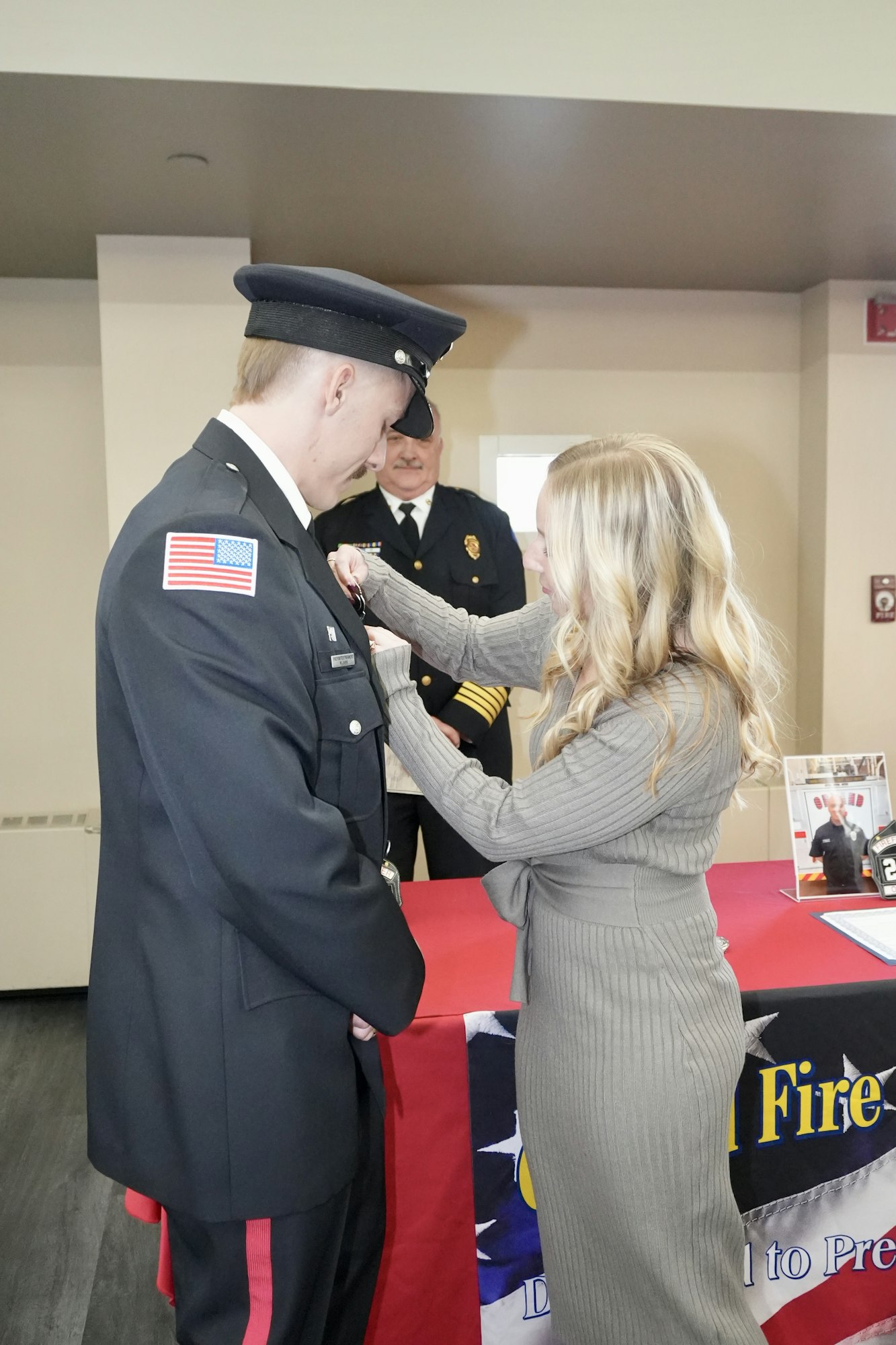A woman pins a badge on a firefighter in uniform during a ceremony, with another uniformed officer in the background.