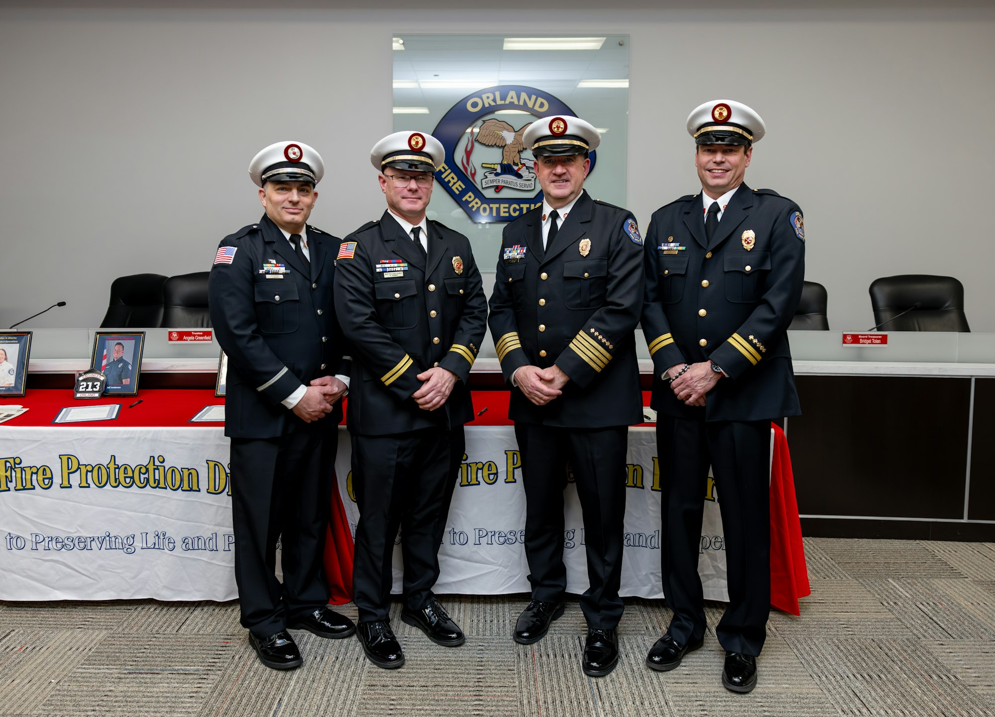Four uniformed fire service officials pose together at a table displaying awards and photographs, promoting fire protection services.