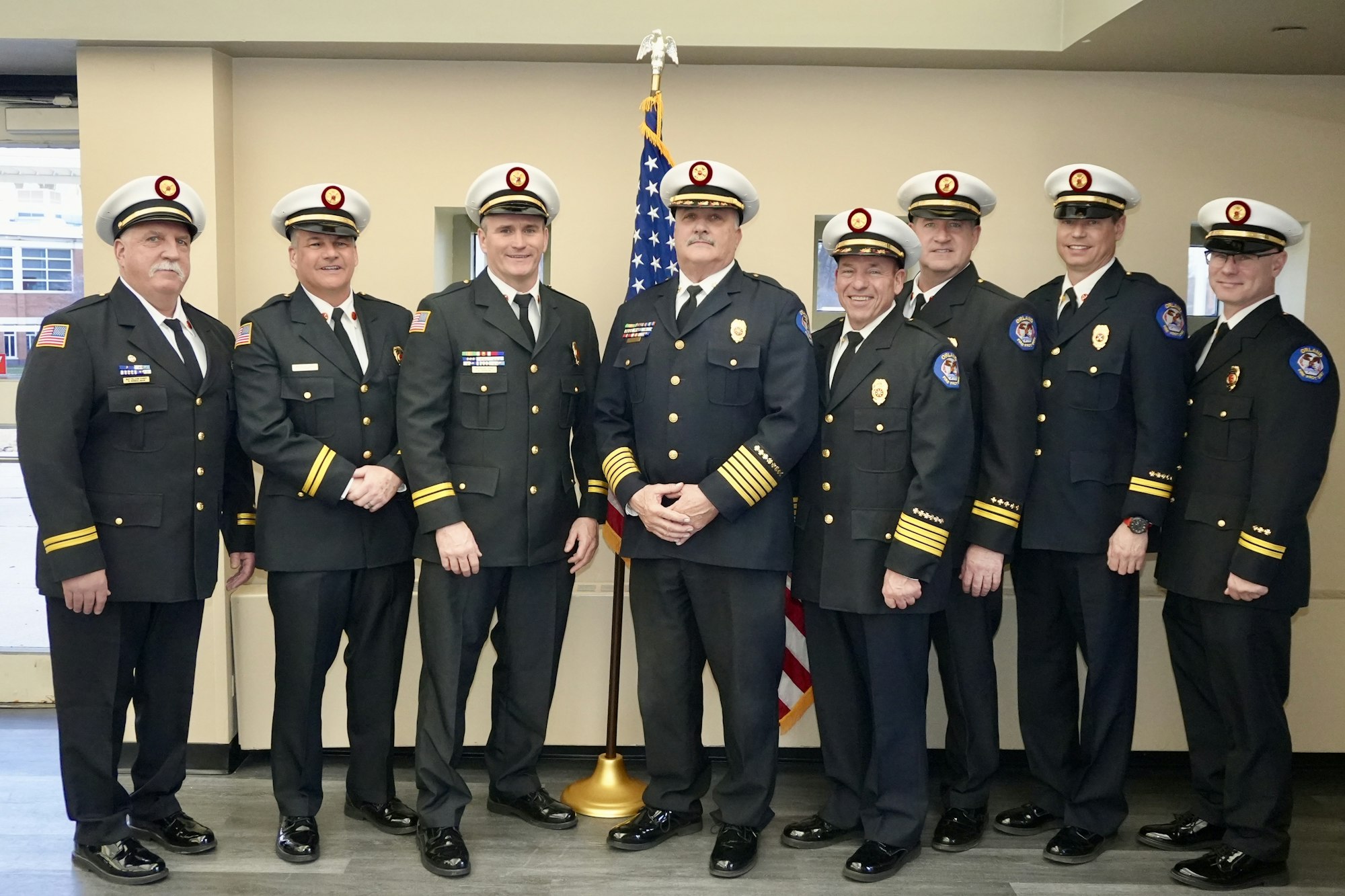 Eight uniformed firefighters pose in front of an American flag indoors.