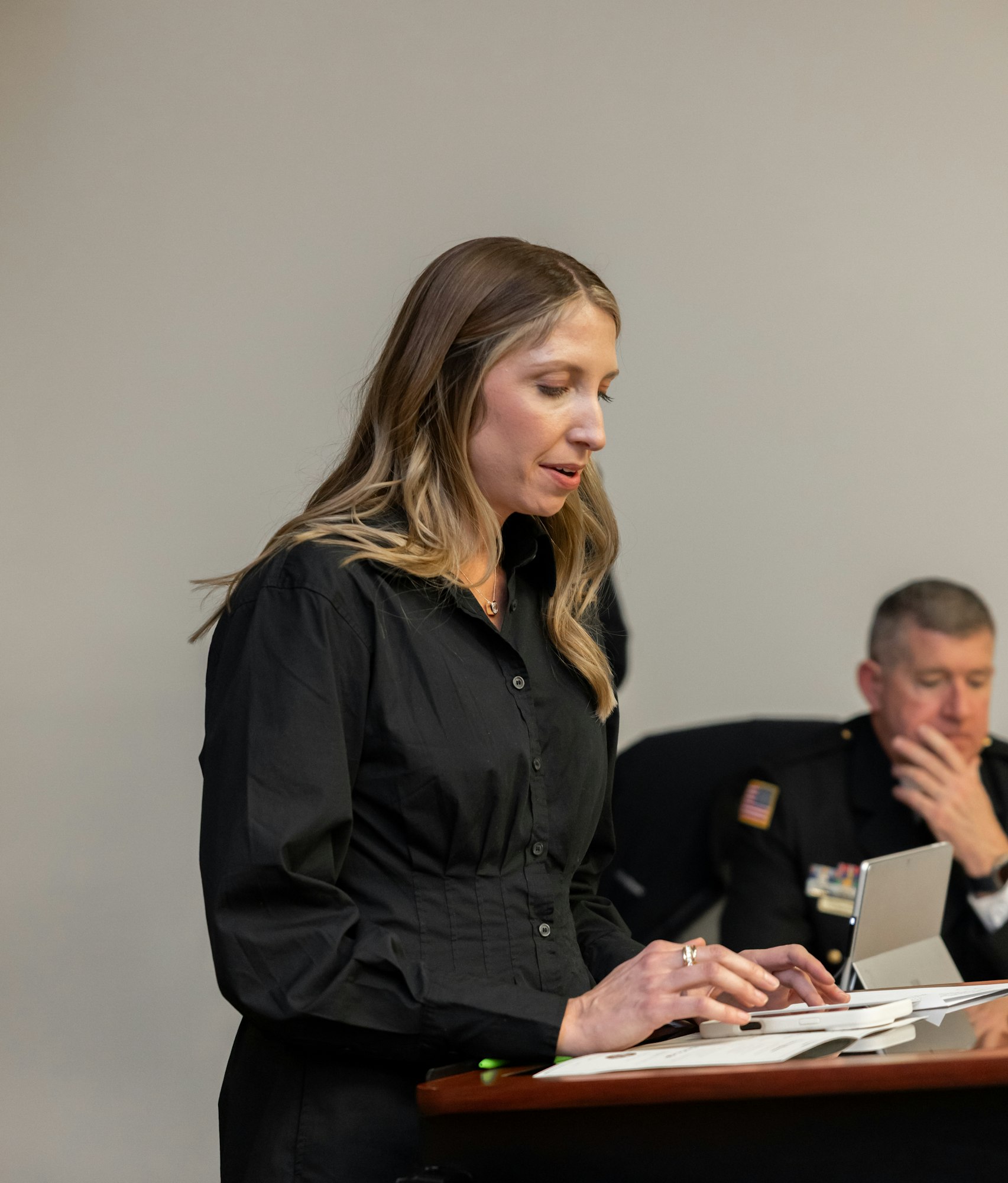 A woman in a black shirt is speaking at a podium, while a man in uniform sits in the background.