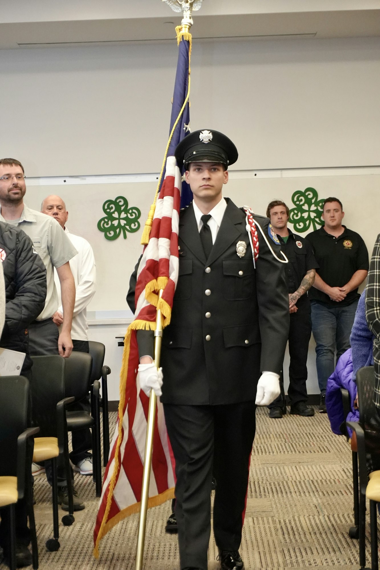 A uniformed man carries an American flag in a ceremony, surrounded by seated and standing attendees.