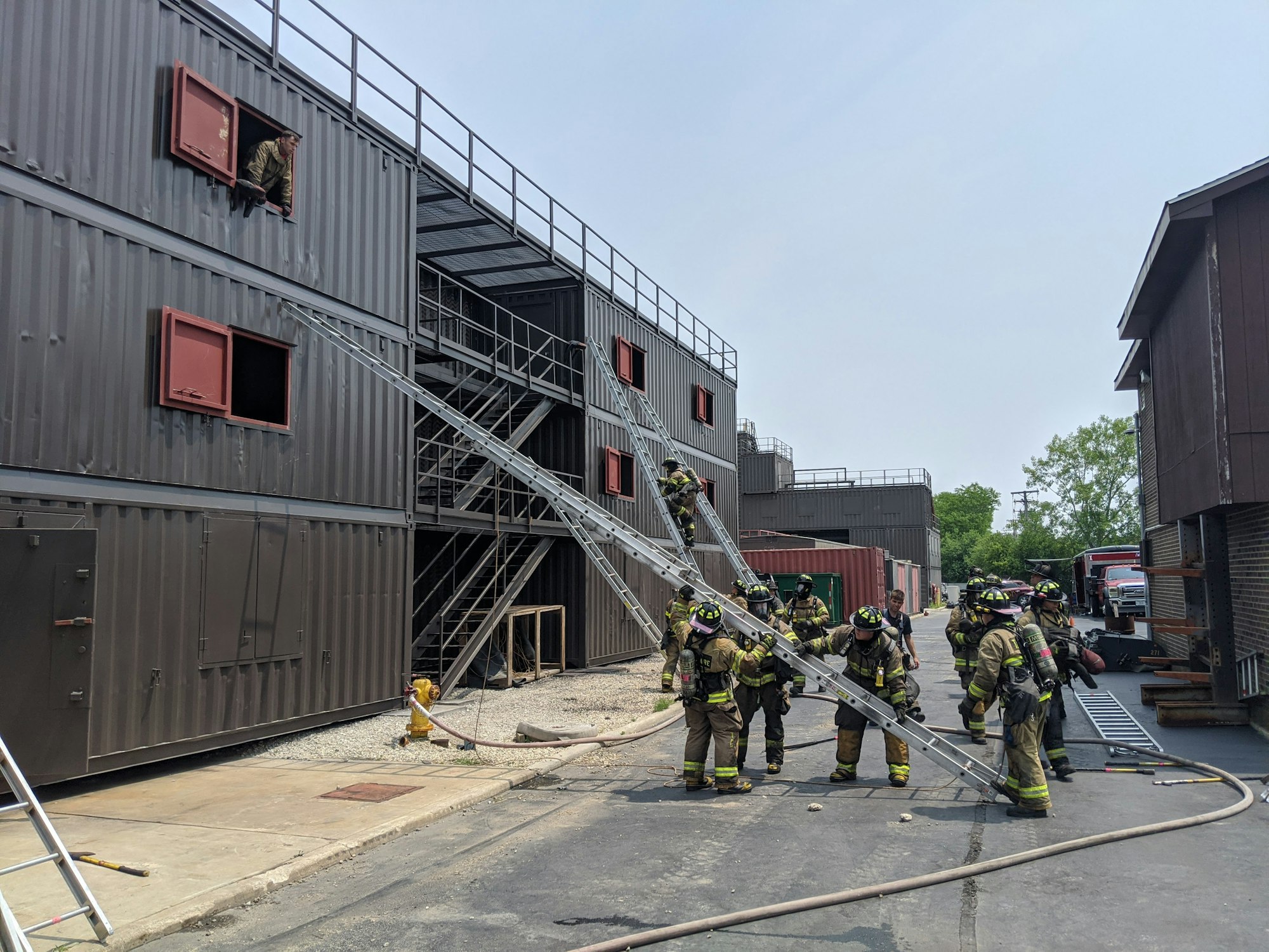 Firefighters training at a fire academy with ladders and hoses on a mock building.