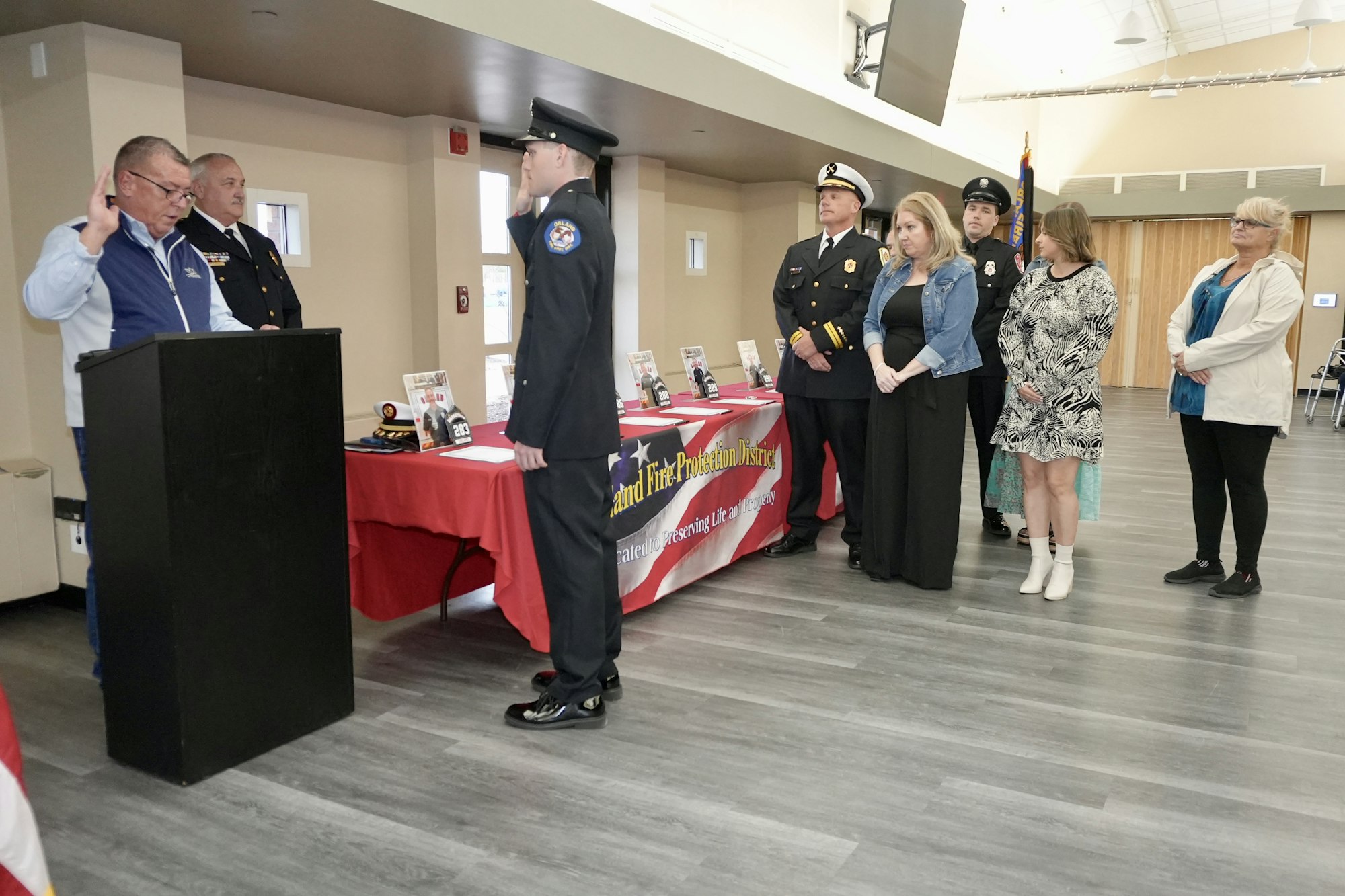 A ceremony with a man swearing in a uniformed officer, observed by others, next to a table with a banner and photos.