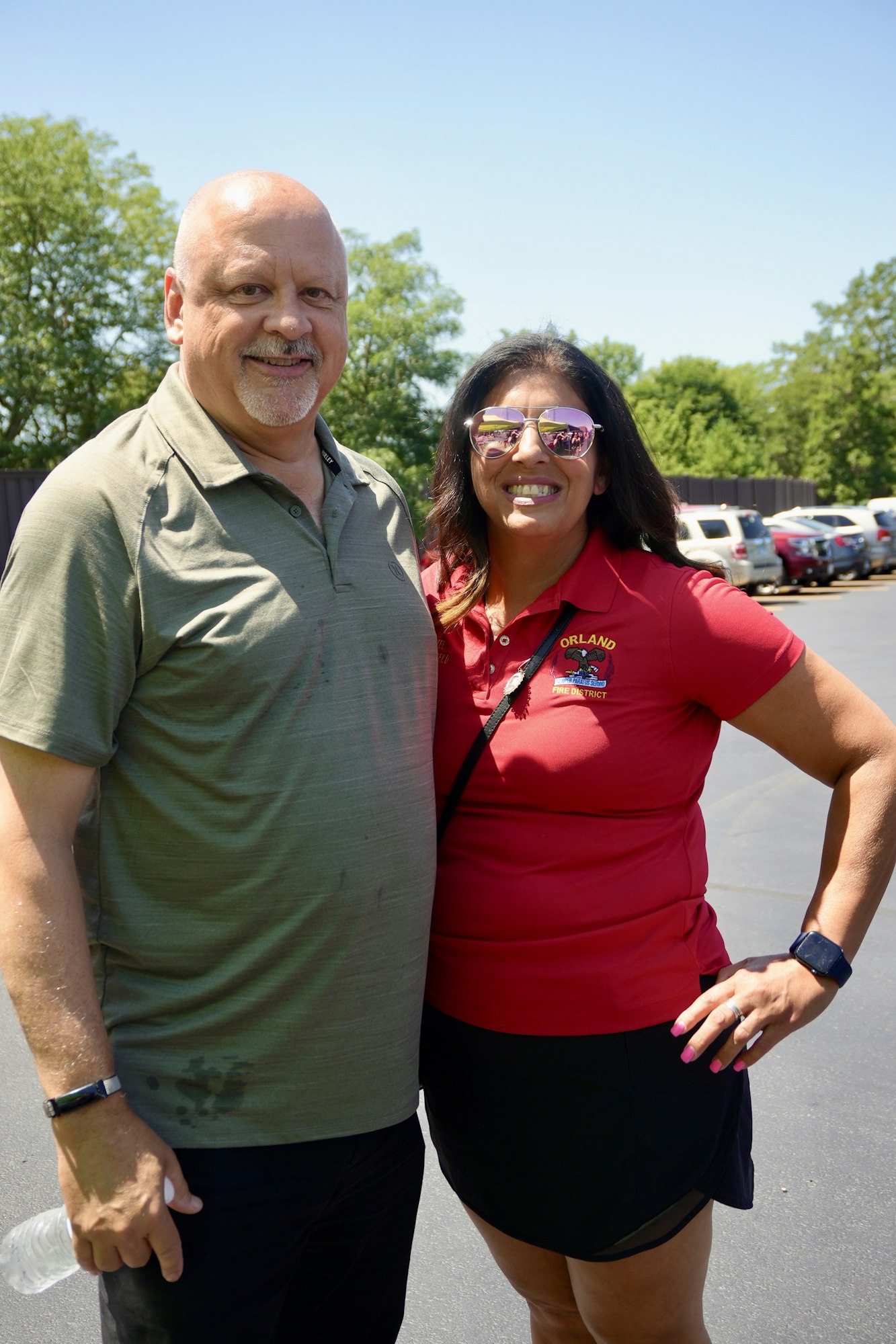 A man and a woman smiling outdoors, with the woman wearing sunglasses and a red shirt that says "Orland."