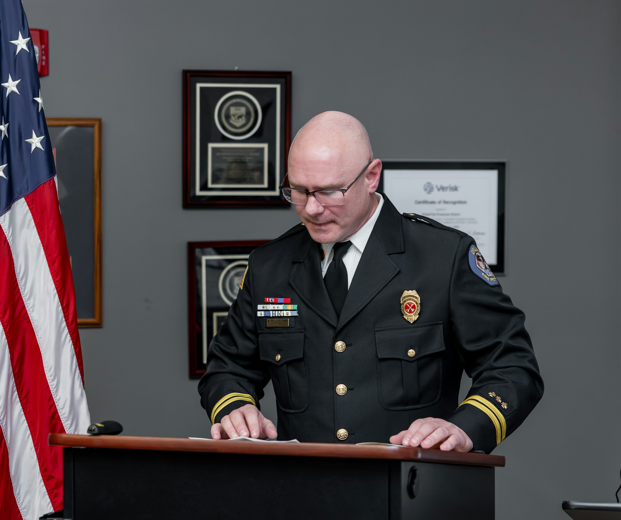 An official in a black uniform stands at a podium, reading documents, with a U.S. flag and framed certificates in the background.