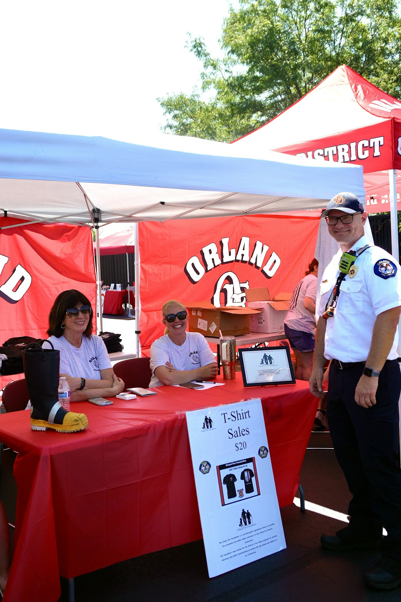 Three people at a booth sell T-shirts for $20. The booth has red "Orland Fire District" banners.