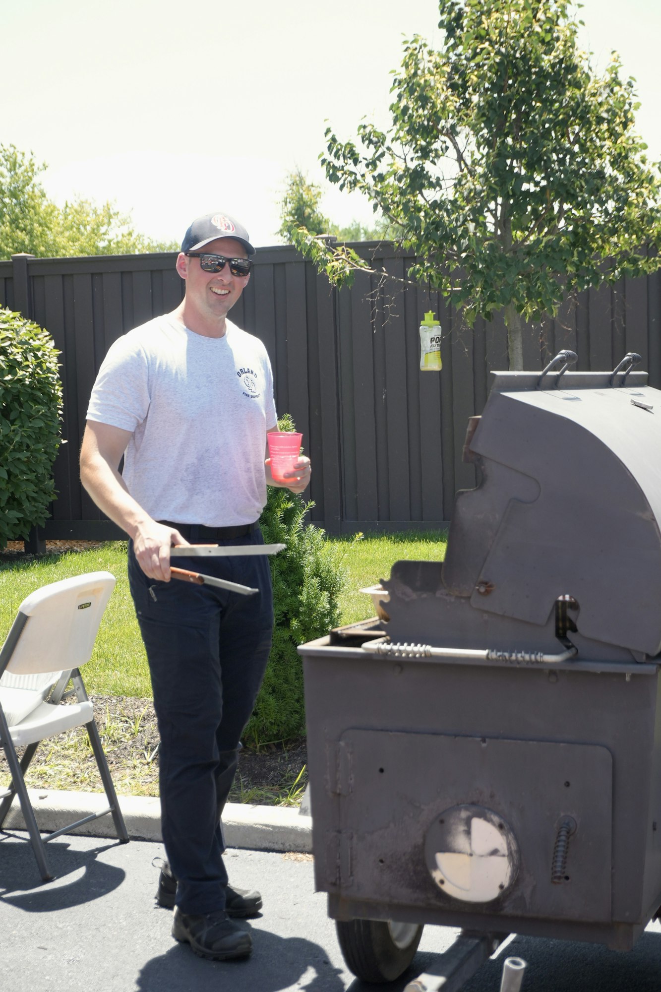 Man grilling outdoors, holding tongs and a cup, wearing a cap and sunglasses, with a grill and a tree in the background.