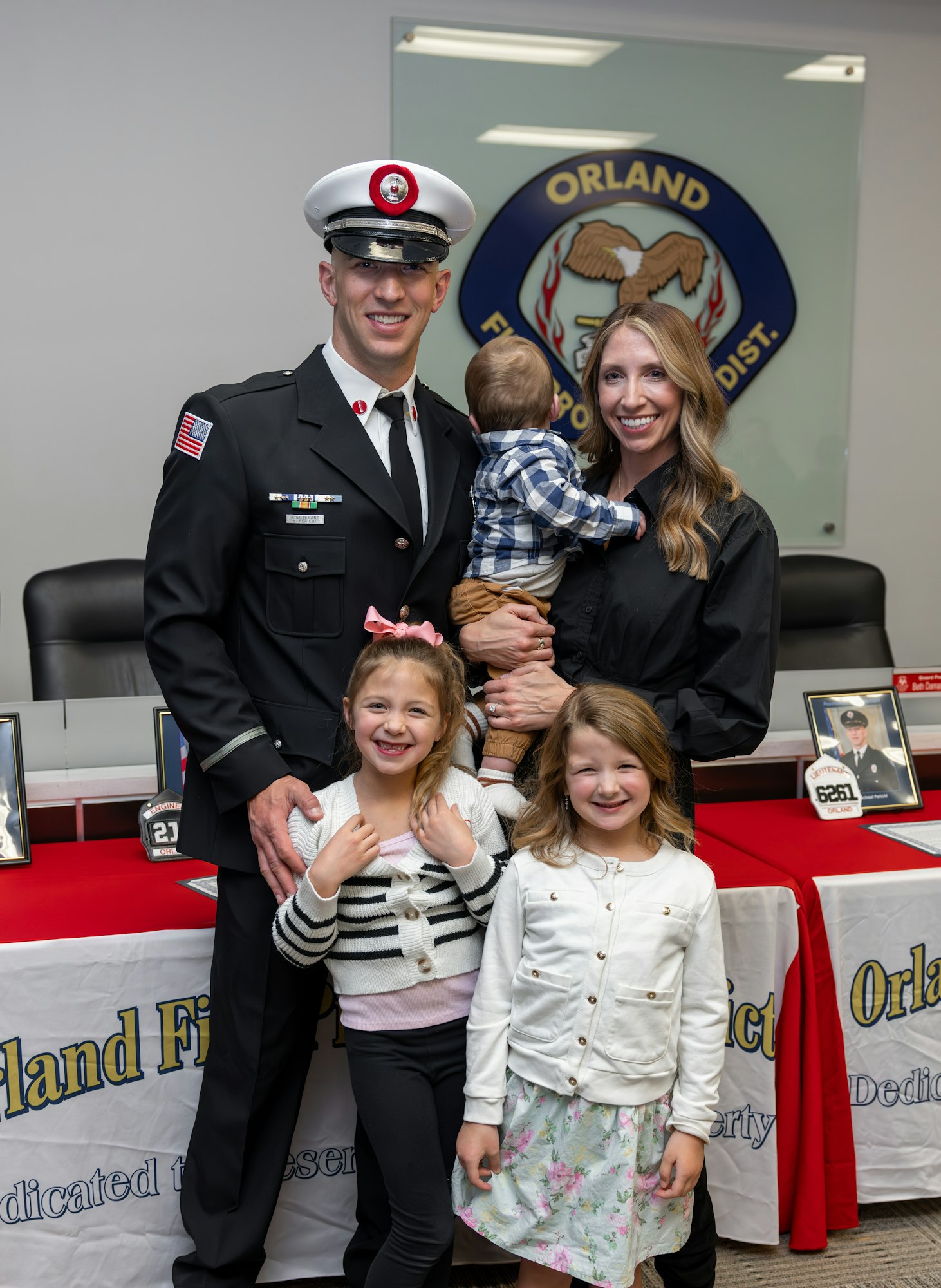 A firefighter in uniform poses with his family, including children and a baby, at a ceremony in an official setting.
