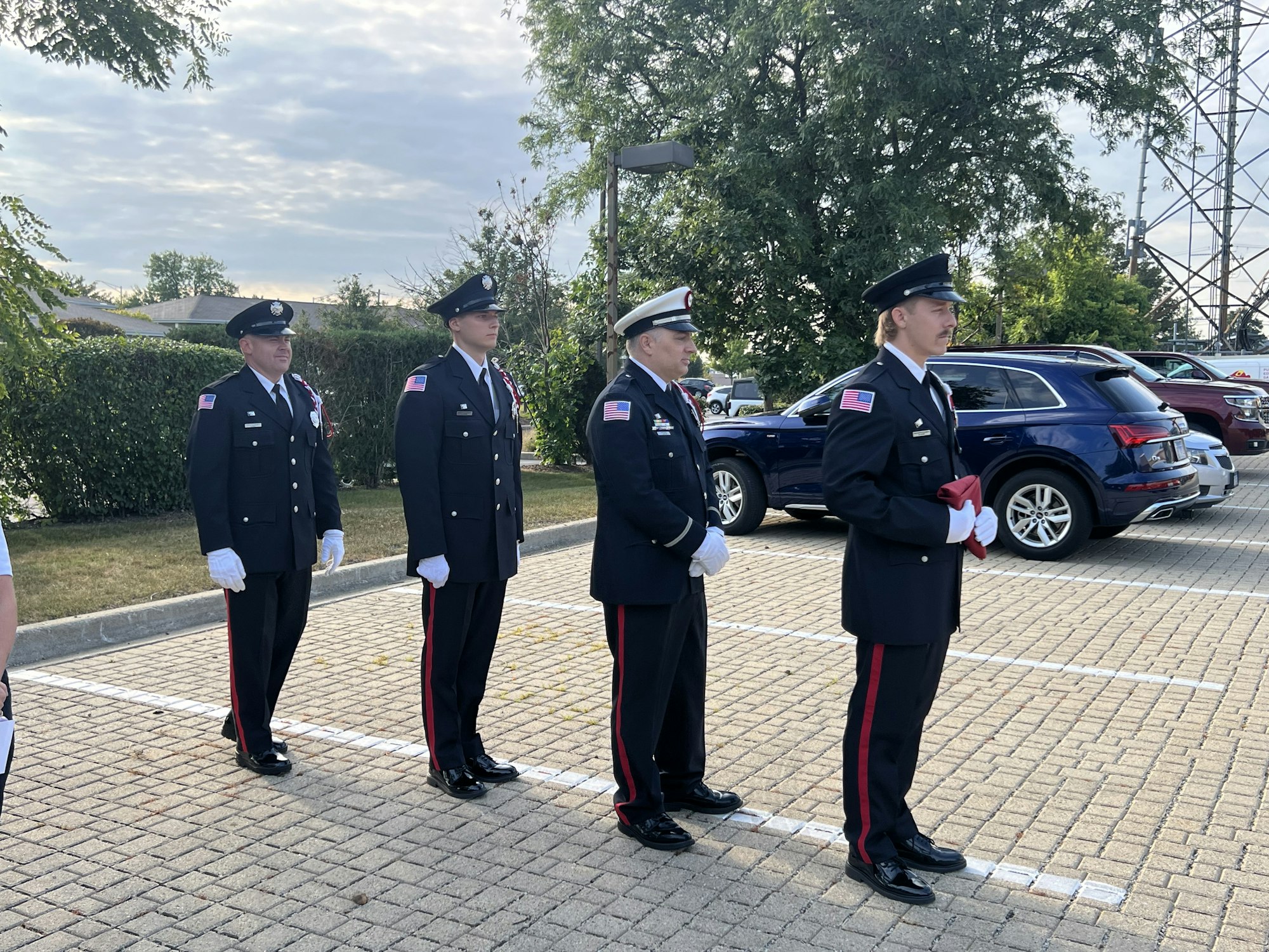 Uniformed individuals standing in formation in a parking lot with parked cars and trees in the background.