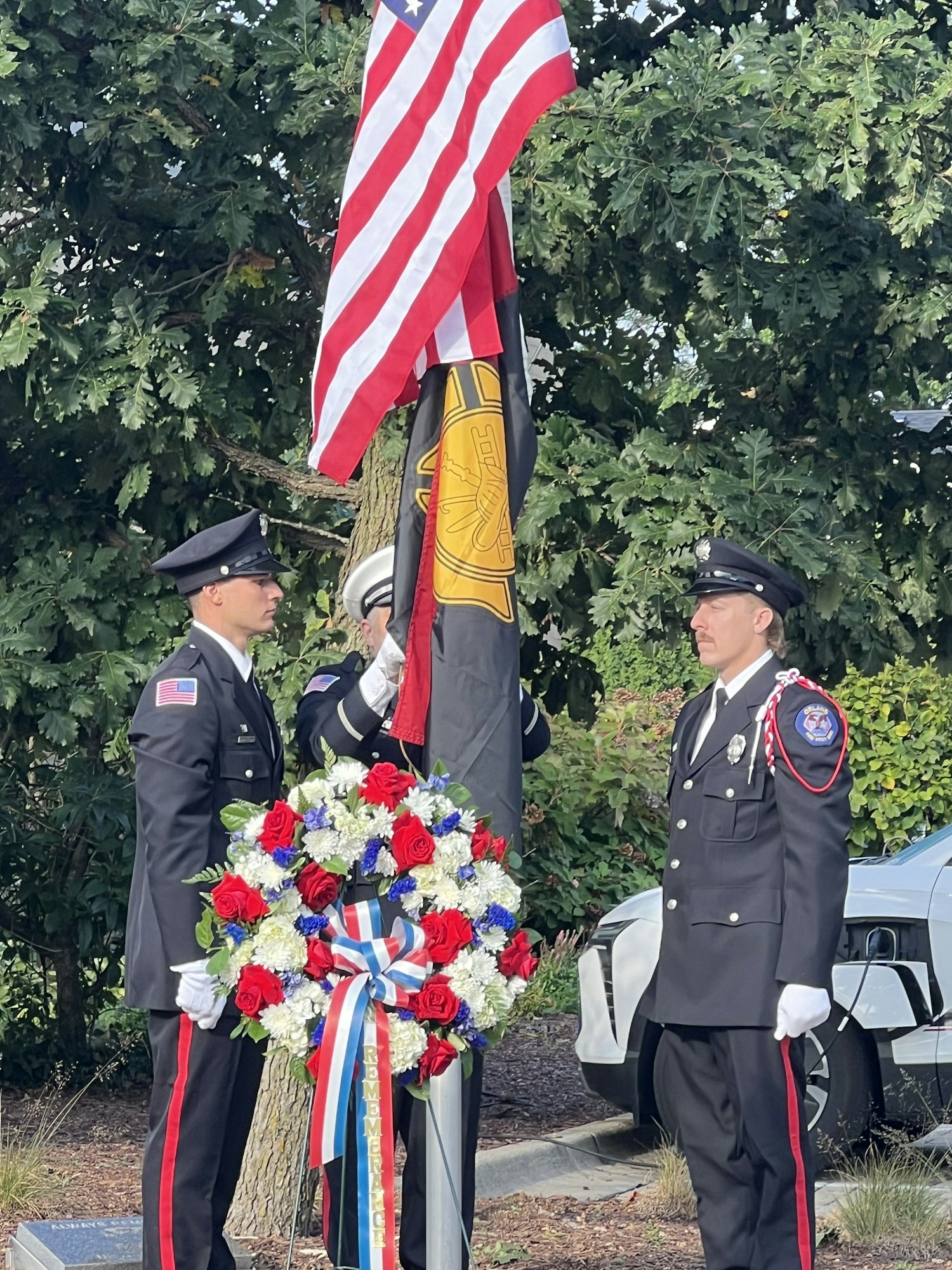 Two uniformed individuals stand by flags and a floral wreath in a ceremonial setting outdoors.