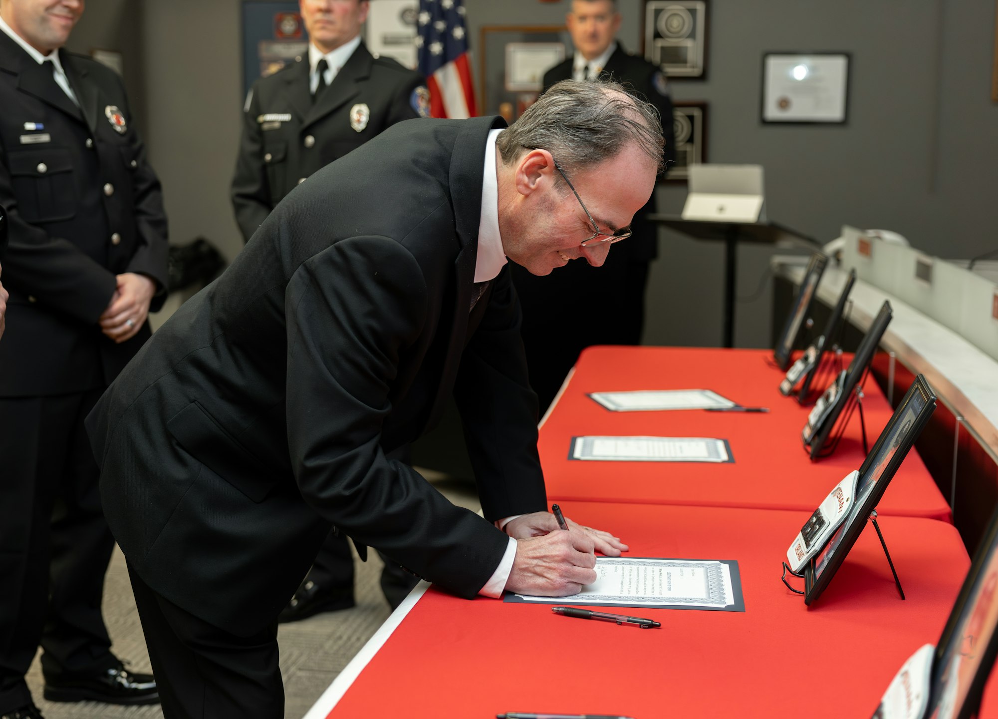 A man in formal attire signs documents at an event, with officers in uniform observing. Red tablecloths are visible.