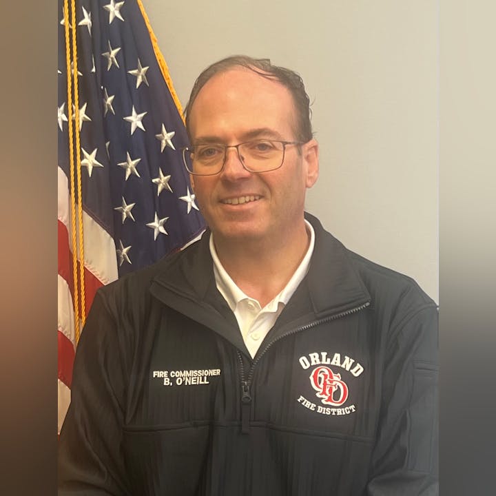 A man in a black jacket with "Orland Fire District" emblem, standing in front of an American flag.