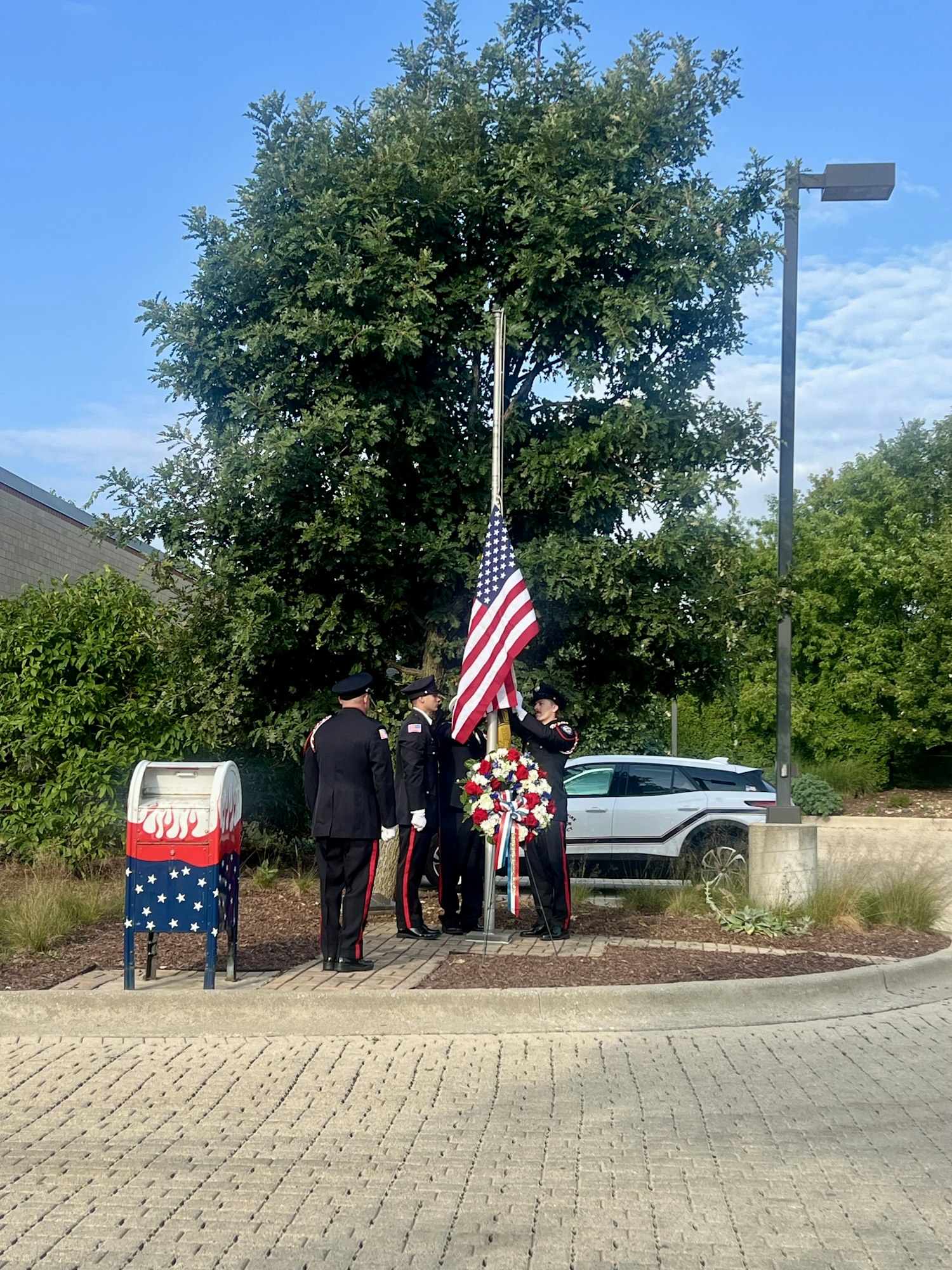 Uniformed individuals holding a U.S. flag at a flagpole with a wreath, near a decorative mailbox.