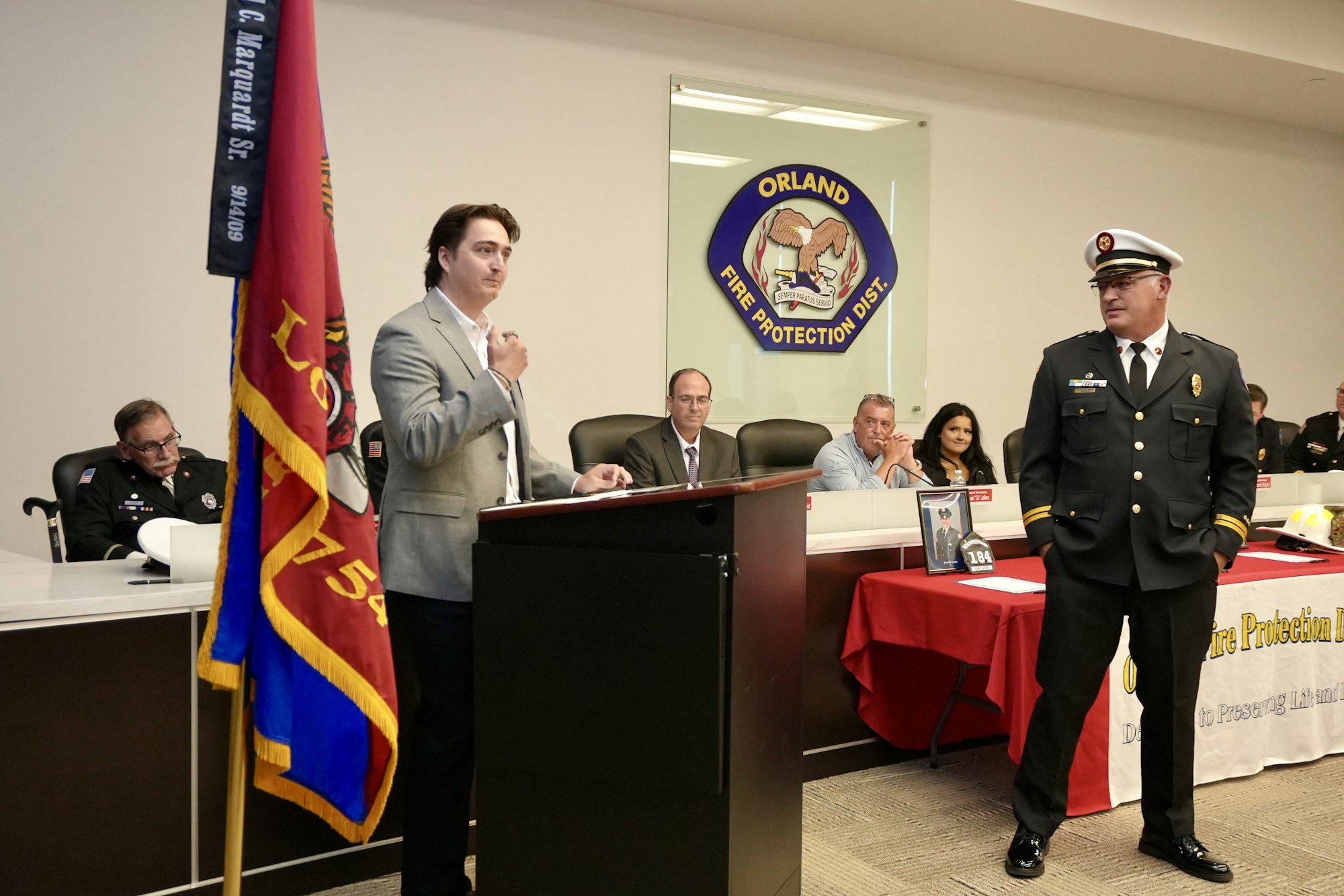 A man speaks at a podium with an Orland Fire Protection District sign behind; others in uniform are seated nearby in a formal setting.