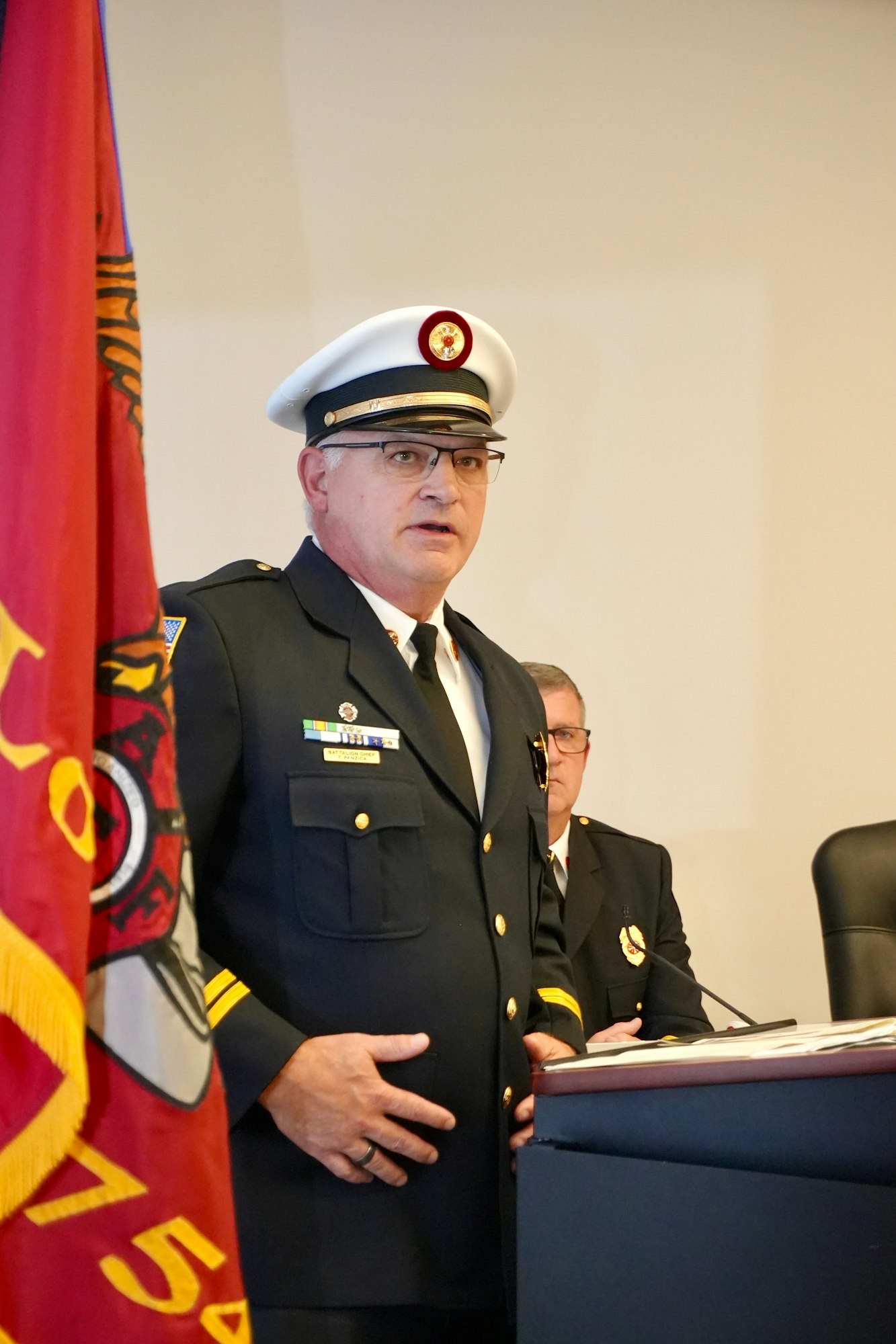 A uniformed fire department official speaking at a podium with a flag in the foreground.