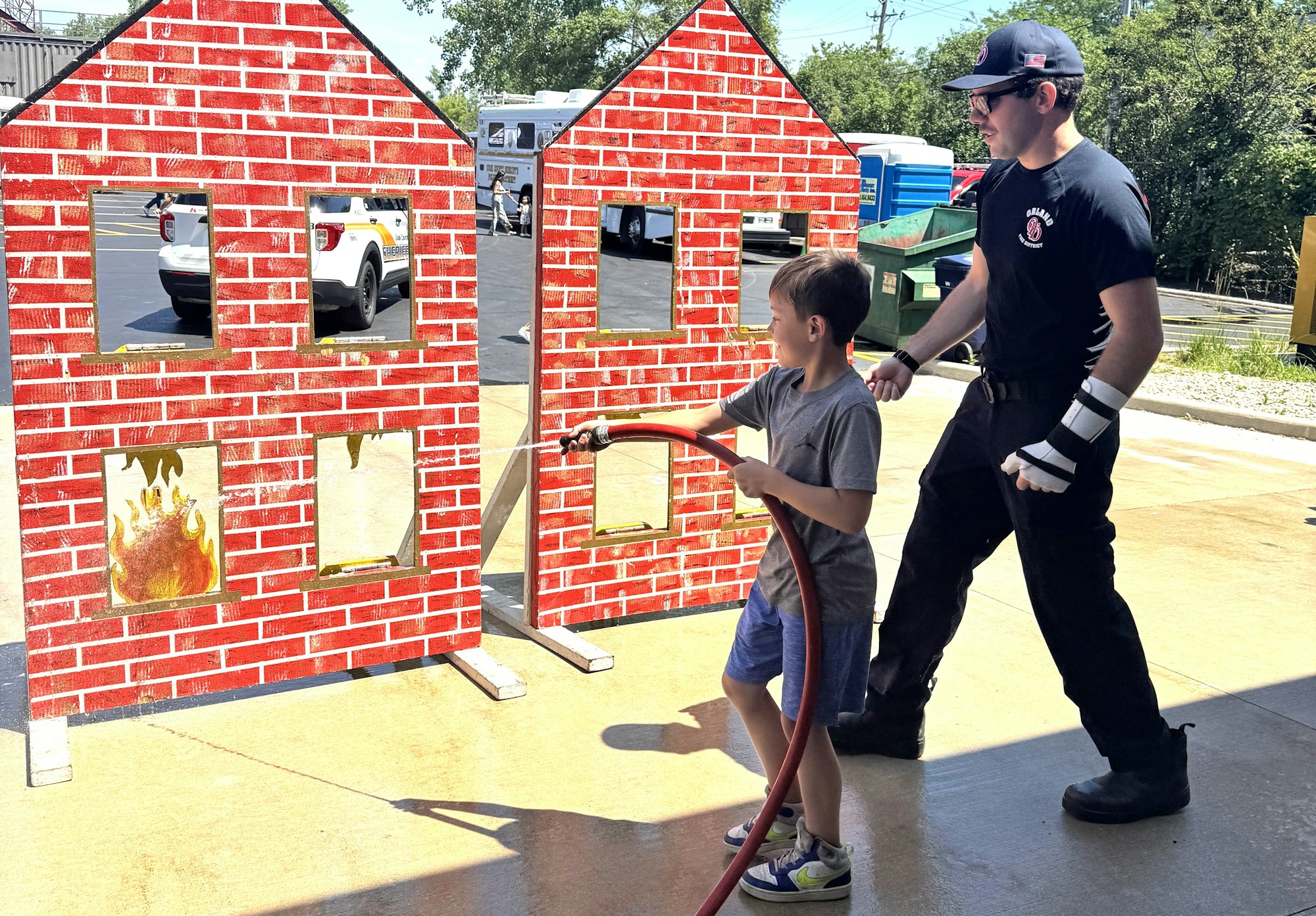 A firefighter assists a child in spraying a hose at a mock-up fire on a wall with painted brick houses.