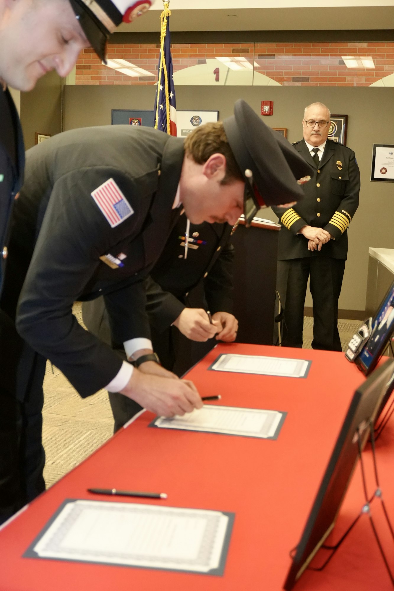 Uniformed individuals signing documents on a red table, with an American flag and framed items in the background.