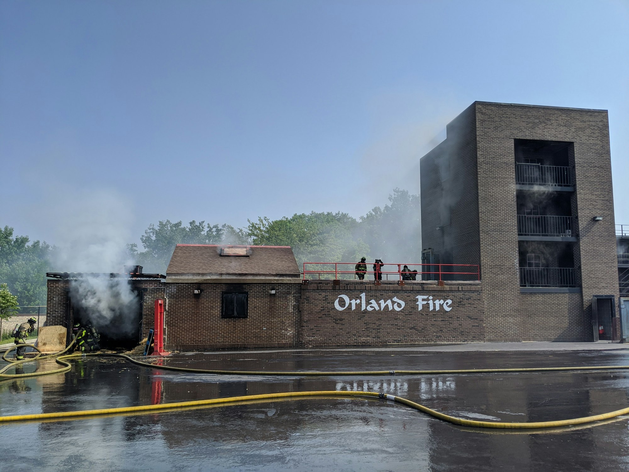 Firefighters extinguishing a fire at a building labeled "Orland Fire." Smoke visible, hoses on ground.
