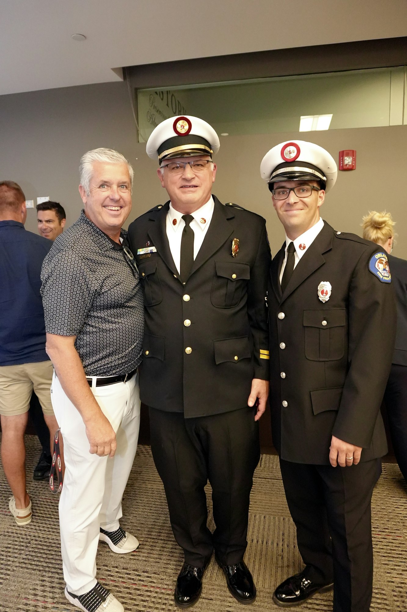 Three men pose together, two in firefighter uniforms and one in casual attire, smiling at the camera.