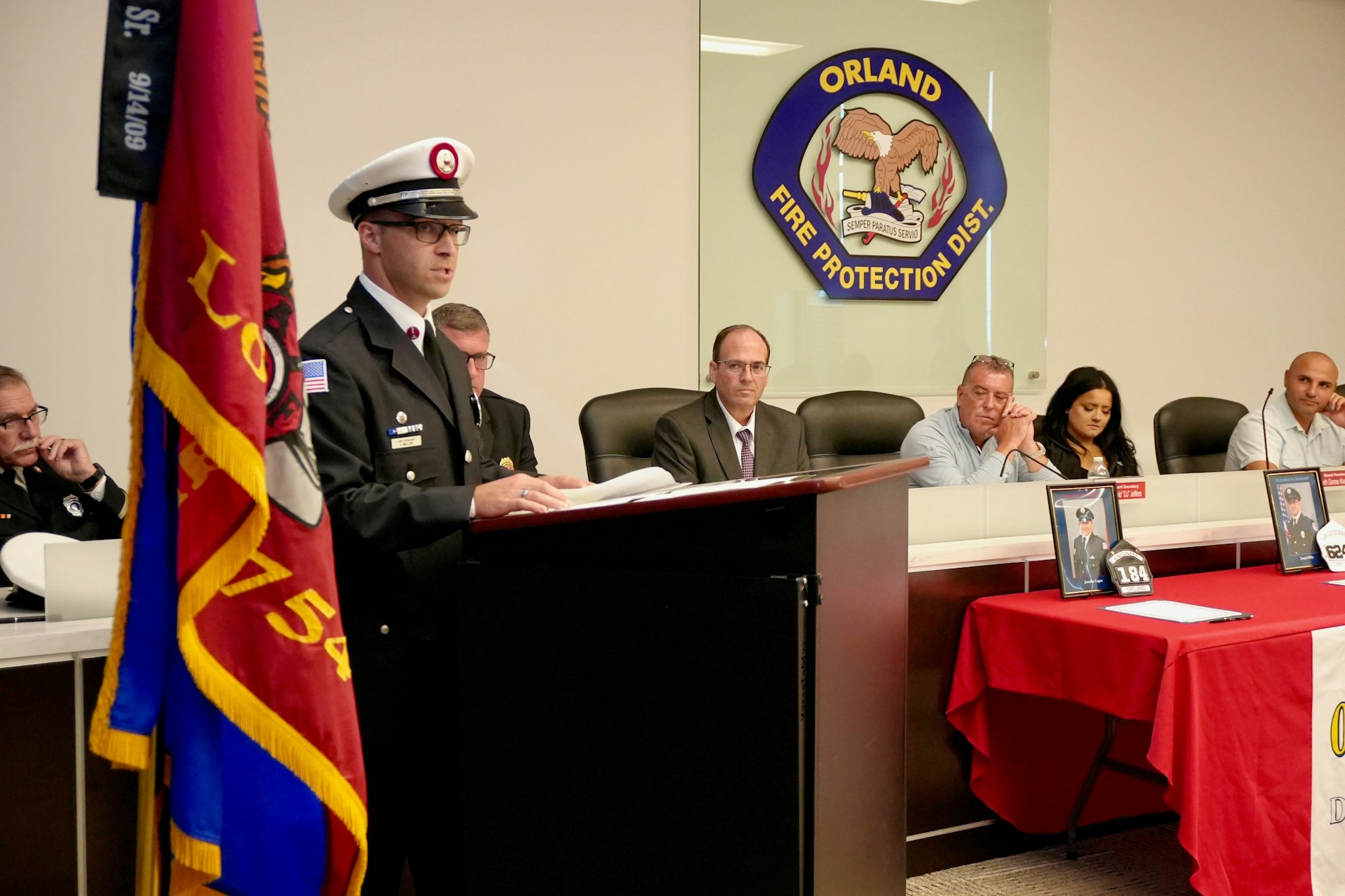 A uniformed speaker at a podium addresses a meeting; the Orland Fire Protection District logo is behind him, and people are seated.