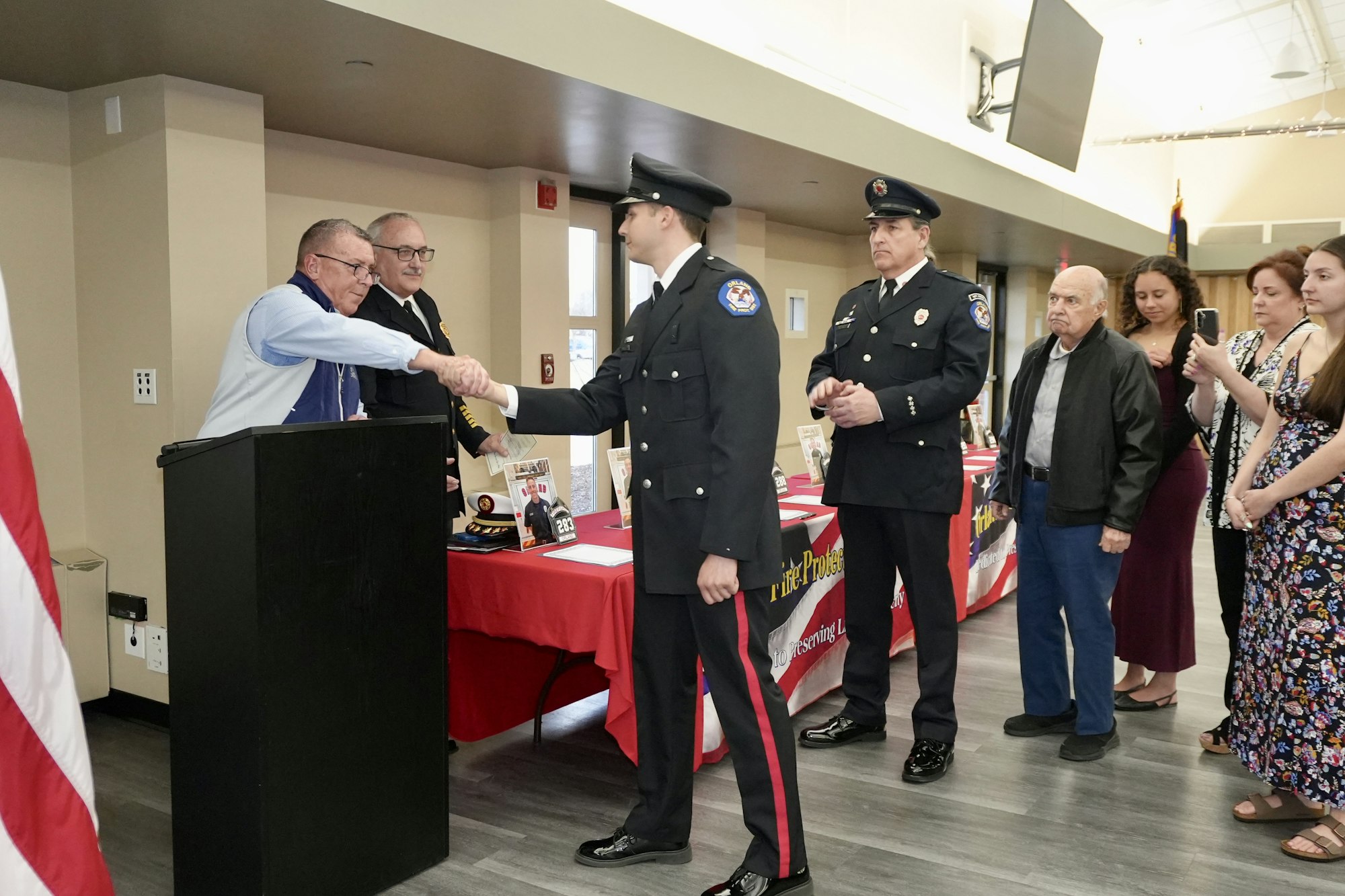 A firefighter in uniform is shaking hands with a man at a ceremony, with other uniformed personnel and spectators nearby.
