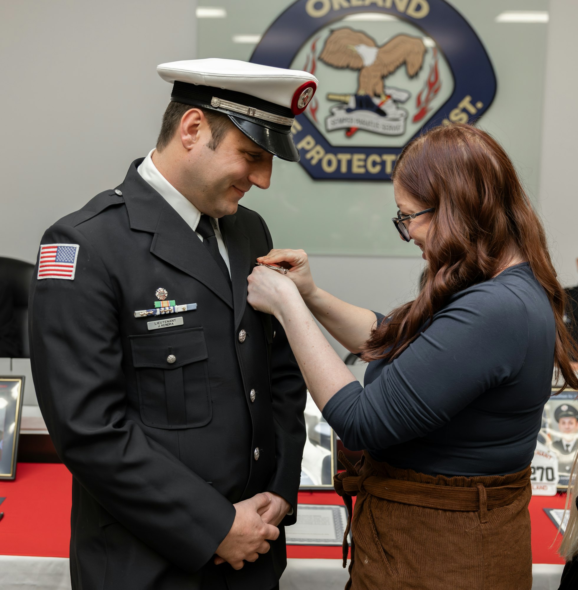 A woman is pinning a badge on a man in uniform, likely during a ceremonial event. Background displays awards and an emblem.