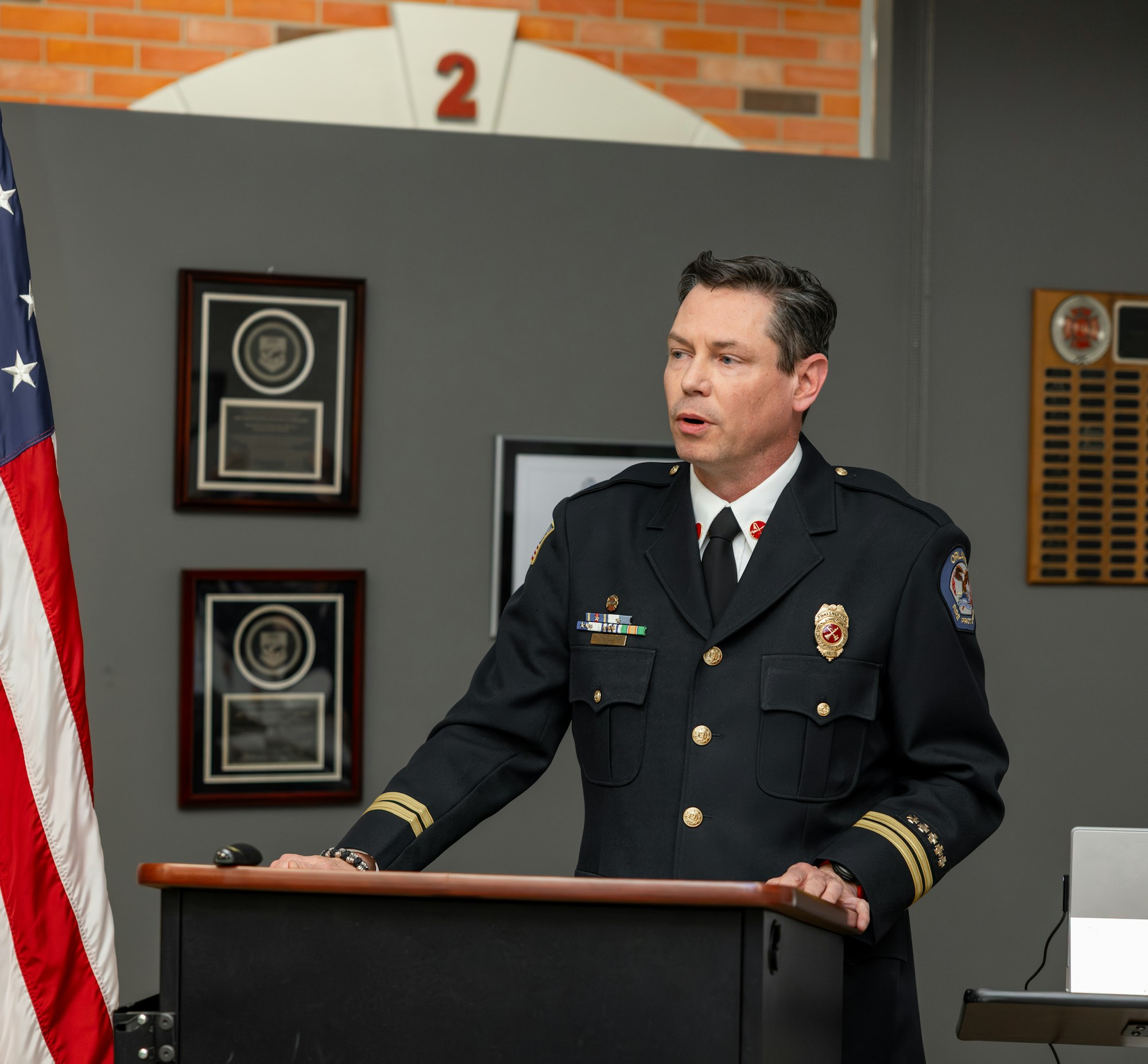 A uniformed officer stands at a podium, speaking, with an American flag and plaques in the background.