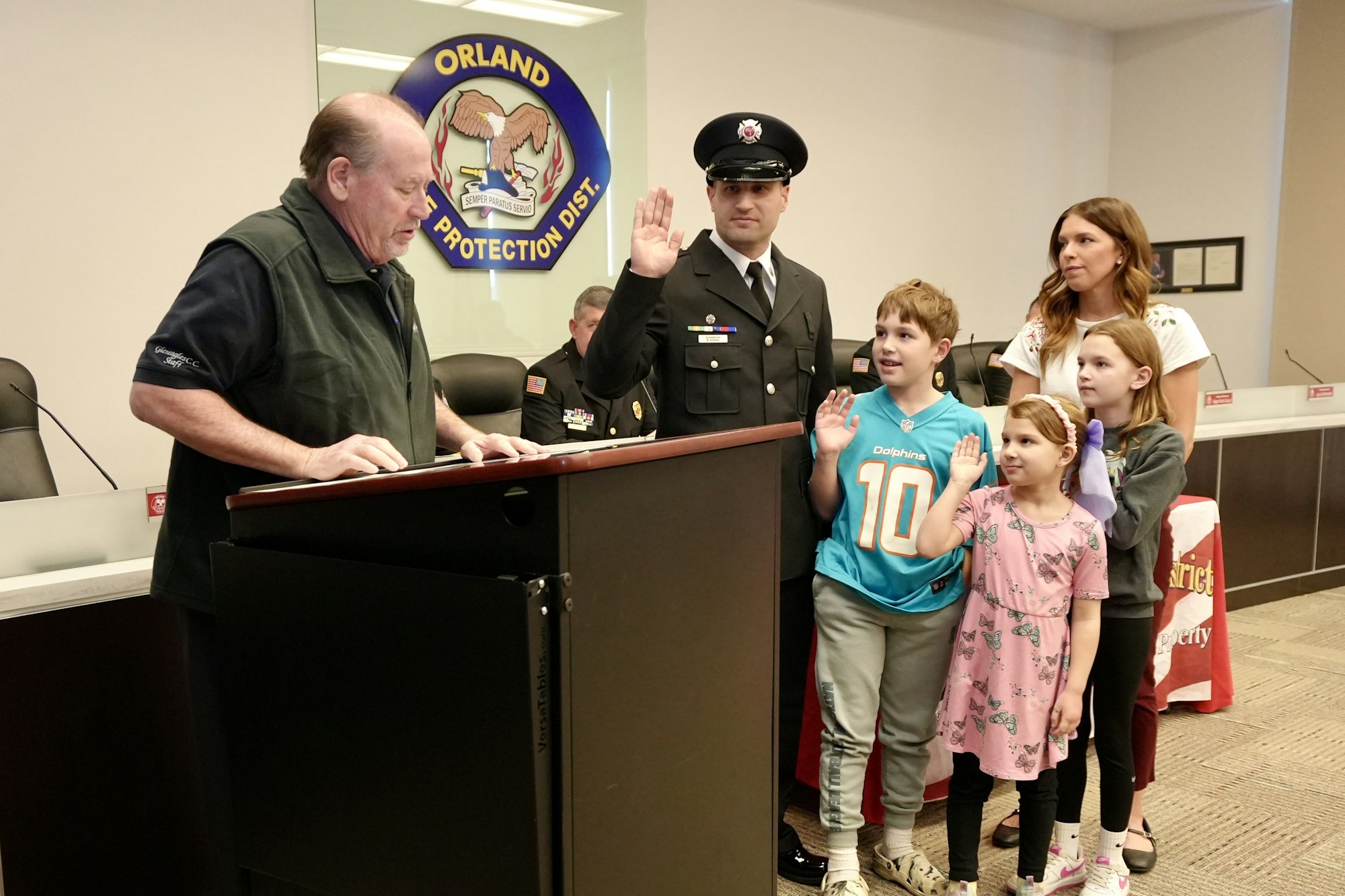 A group of people, including a uniformed man and three kids, participate in an oath ceremony at a podium, with "Orland Fire Protection" in the background.