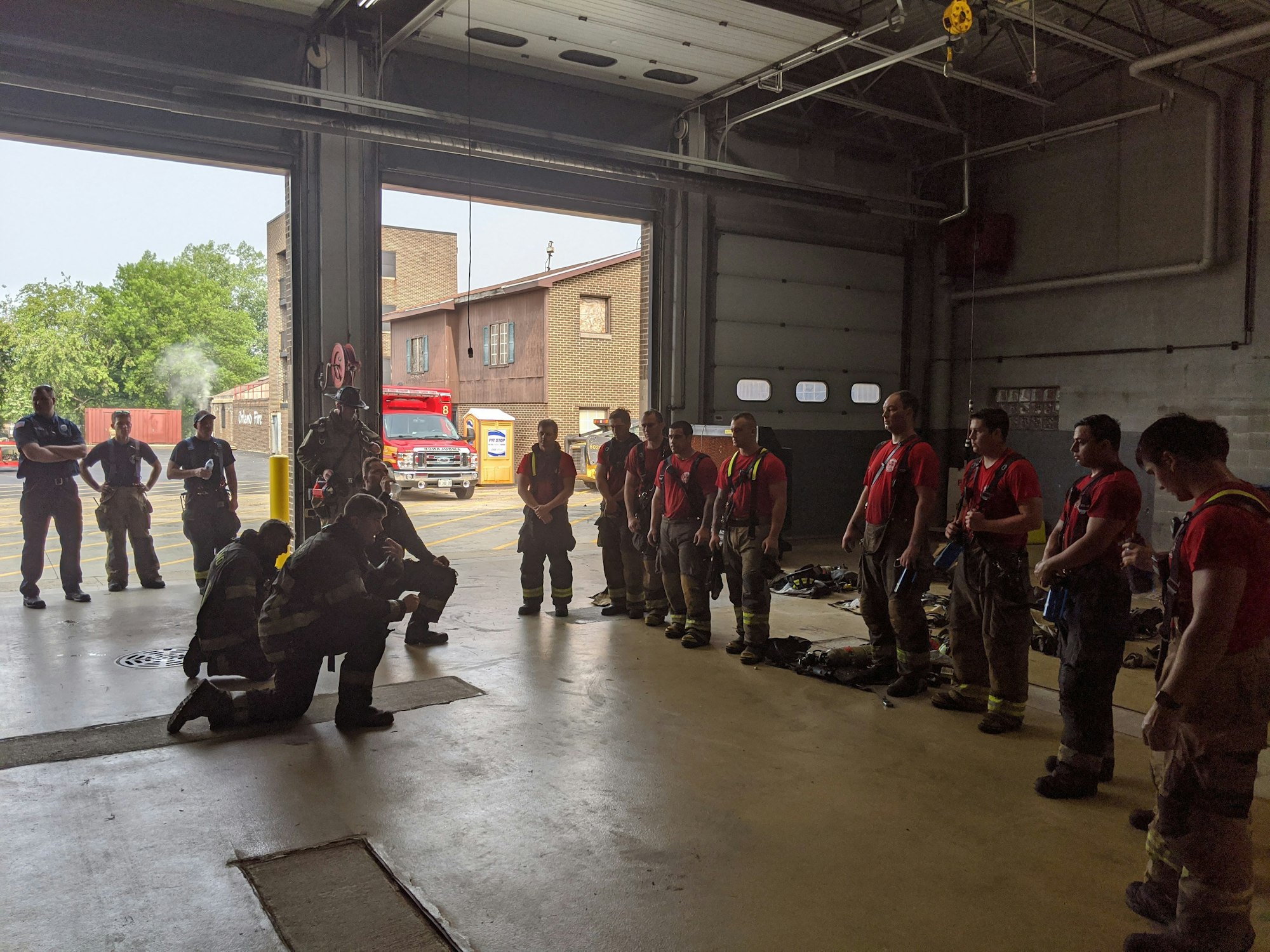 Firefighters gathered in a garage bay, some standing, others sitting, with equipment on the ground and a fire truck visible outside.
