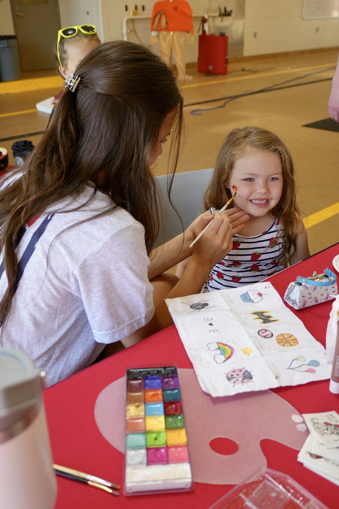 A child getting face paint, smiling, with colorful face paint palette and design samples on the table.