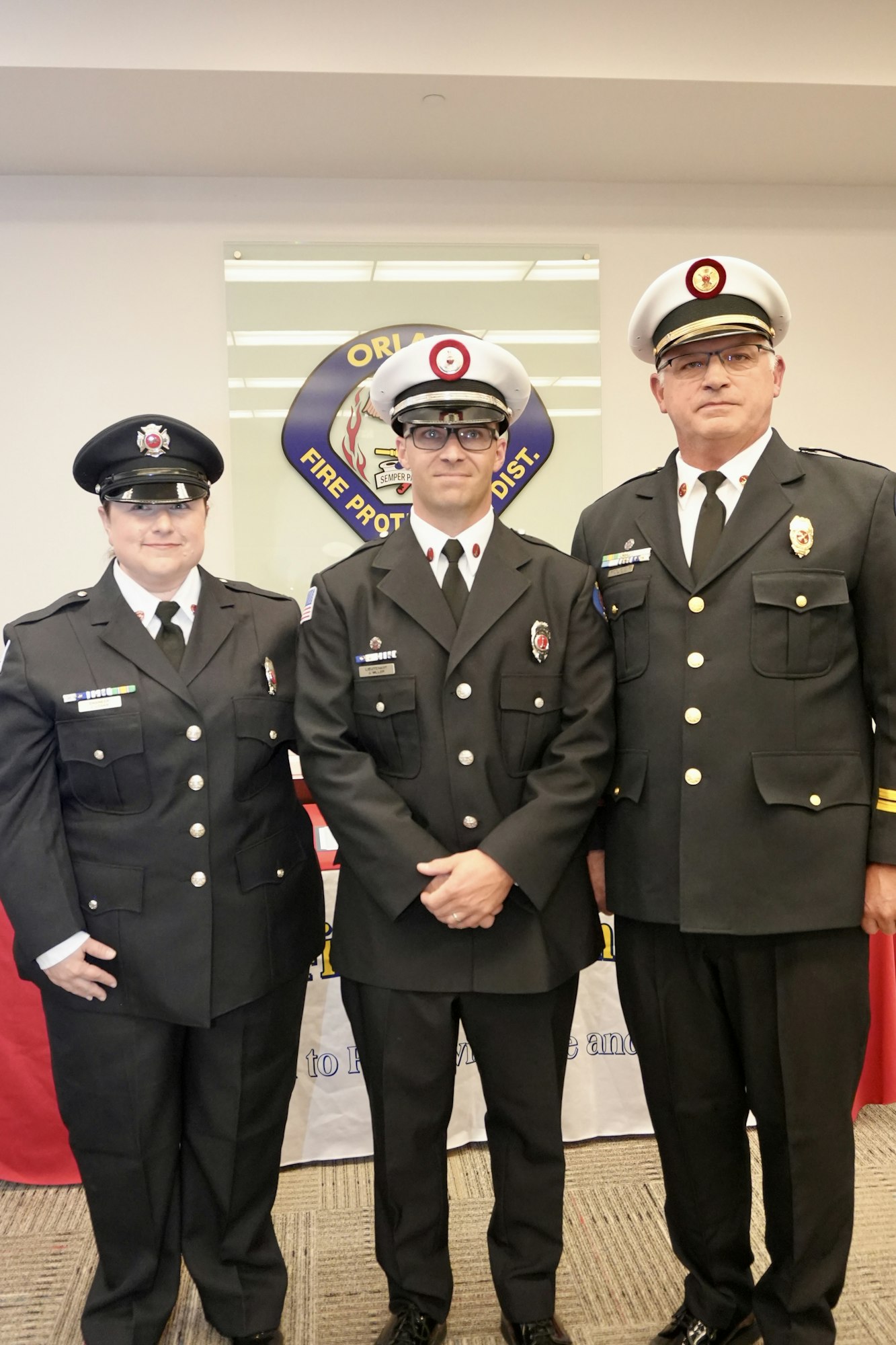 Three people in firefighter uniforms stand in front of a "Fire Protection Dist." sign.