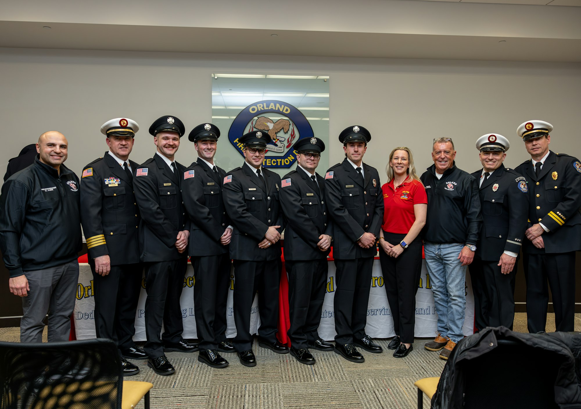 A group of firefighters and officials in uniform pose for a photo at an event in front of an "Orland Fire Protection" banner.