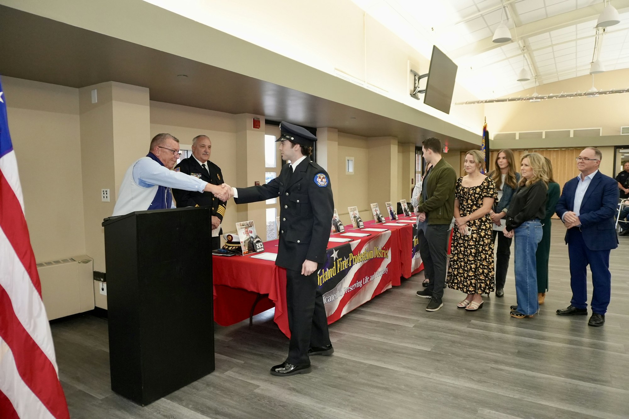 A person in uniform shakes hands with an official at a ceremony, with a group of people watching beside a table with brochures.
