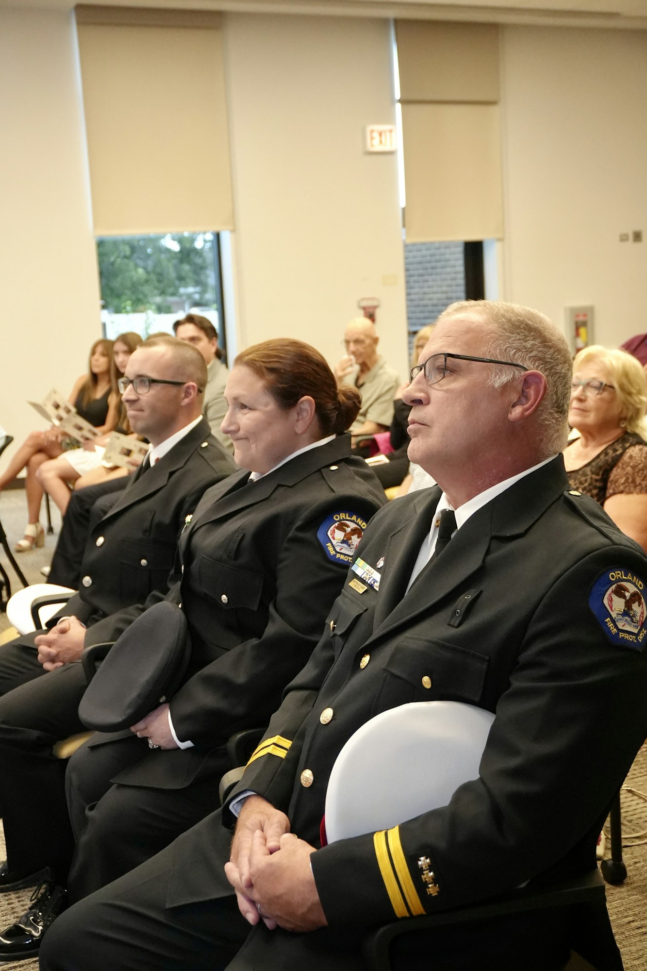 Uniformed officers seated in a row at an indoor event, with other attendees in the background.
