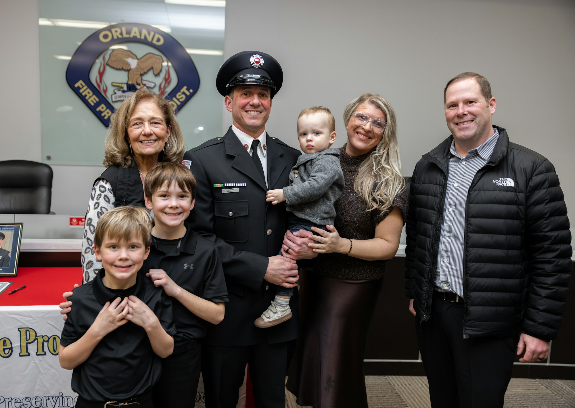 A group of seven people, including children and an adult in a fire uniform, pose together in front of a fire department backdrop.