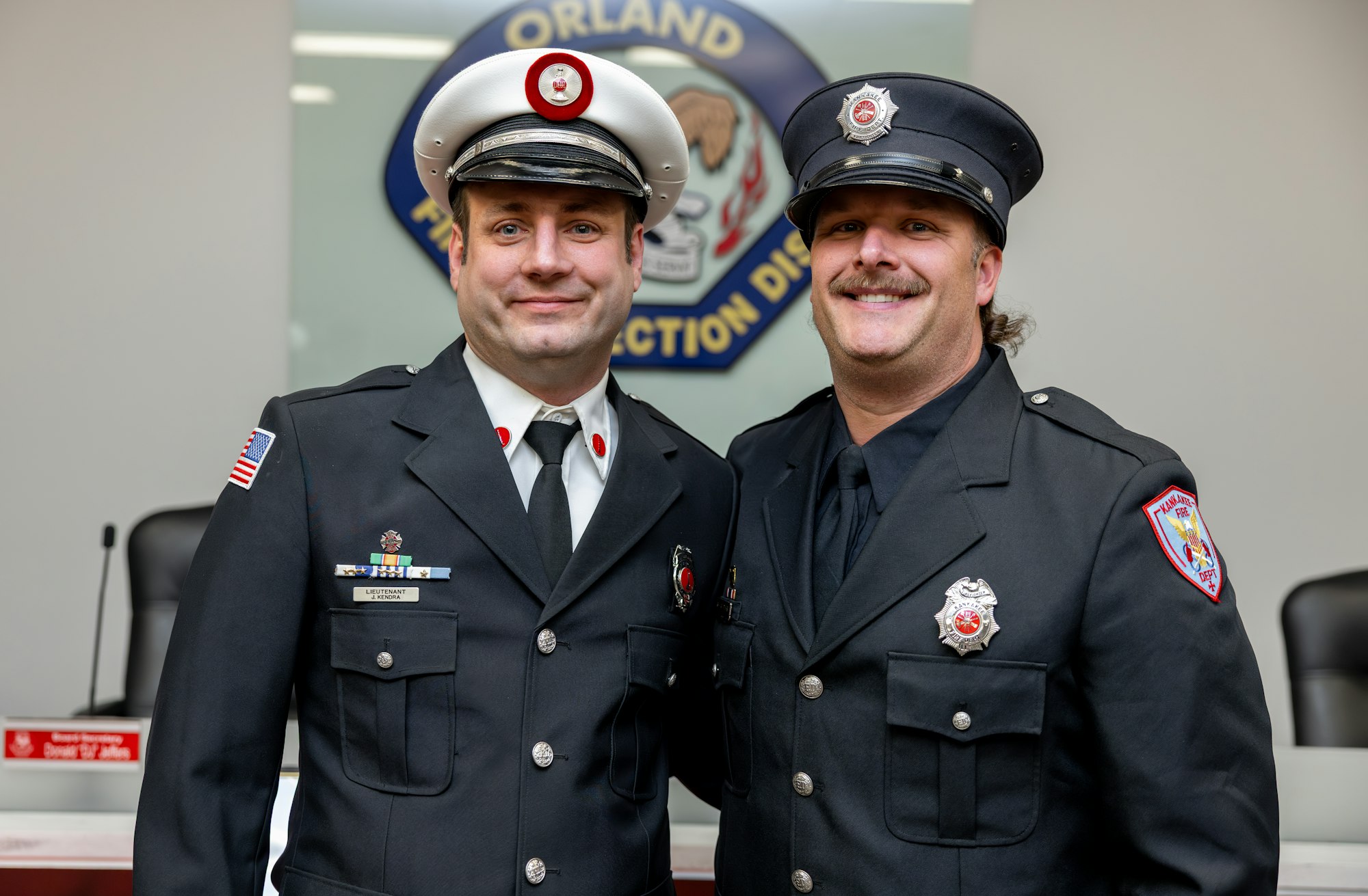 The image shows two firefighters in uniforms, smiling, with a badge and an official logo in the background.